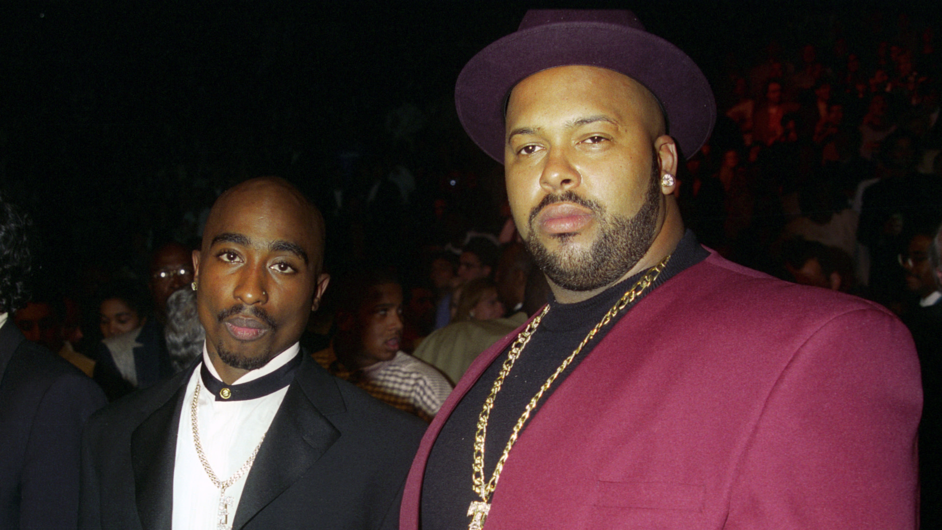Tupac Shakur and Suge Knight at an event. Tupac is in a black suit, and Suge is wearing a maroon suit and hat with gold jewelry.