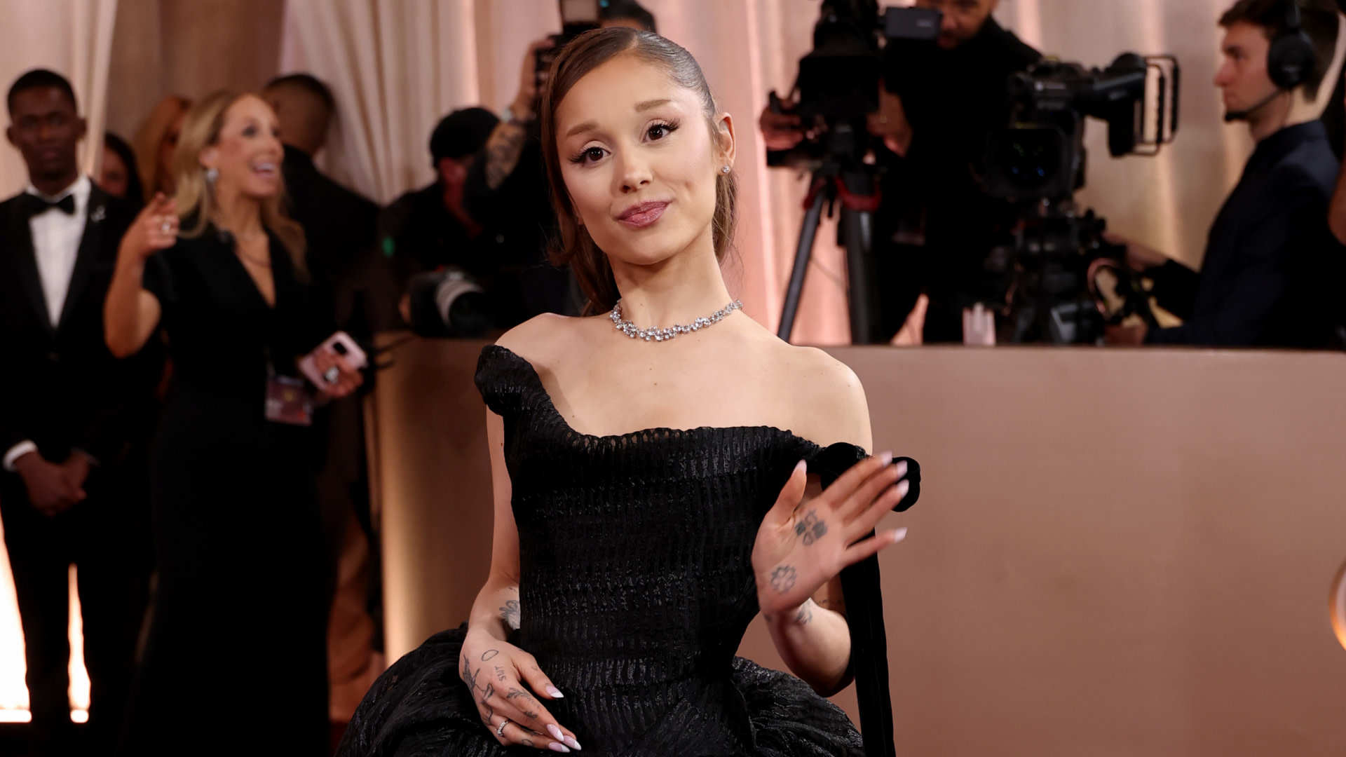 Ariana Grande waves on a red carpet, wearing a black dress and necklace, with cameras and people in the background.