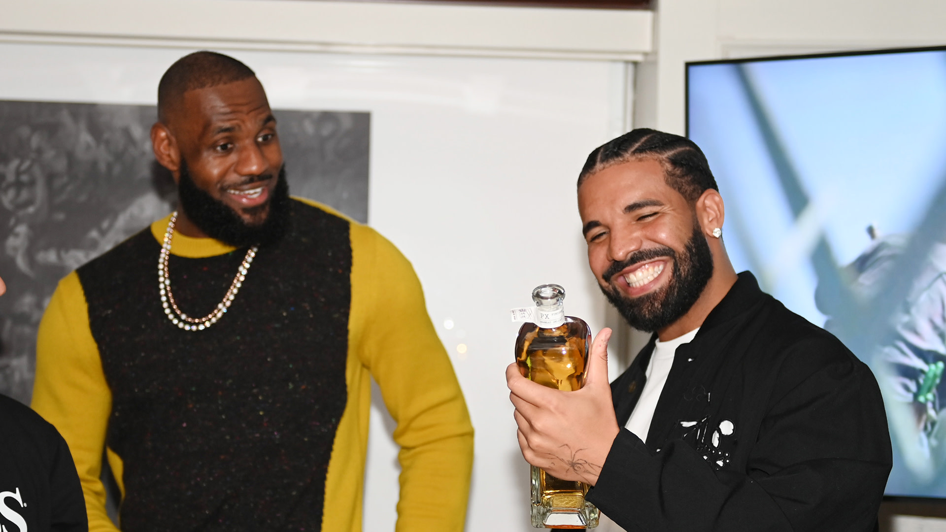 LeBron James in a yellow sweater looks at Drake, who is smiling and holding a bottle of liquor.
