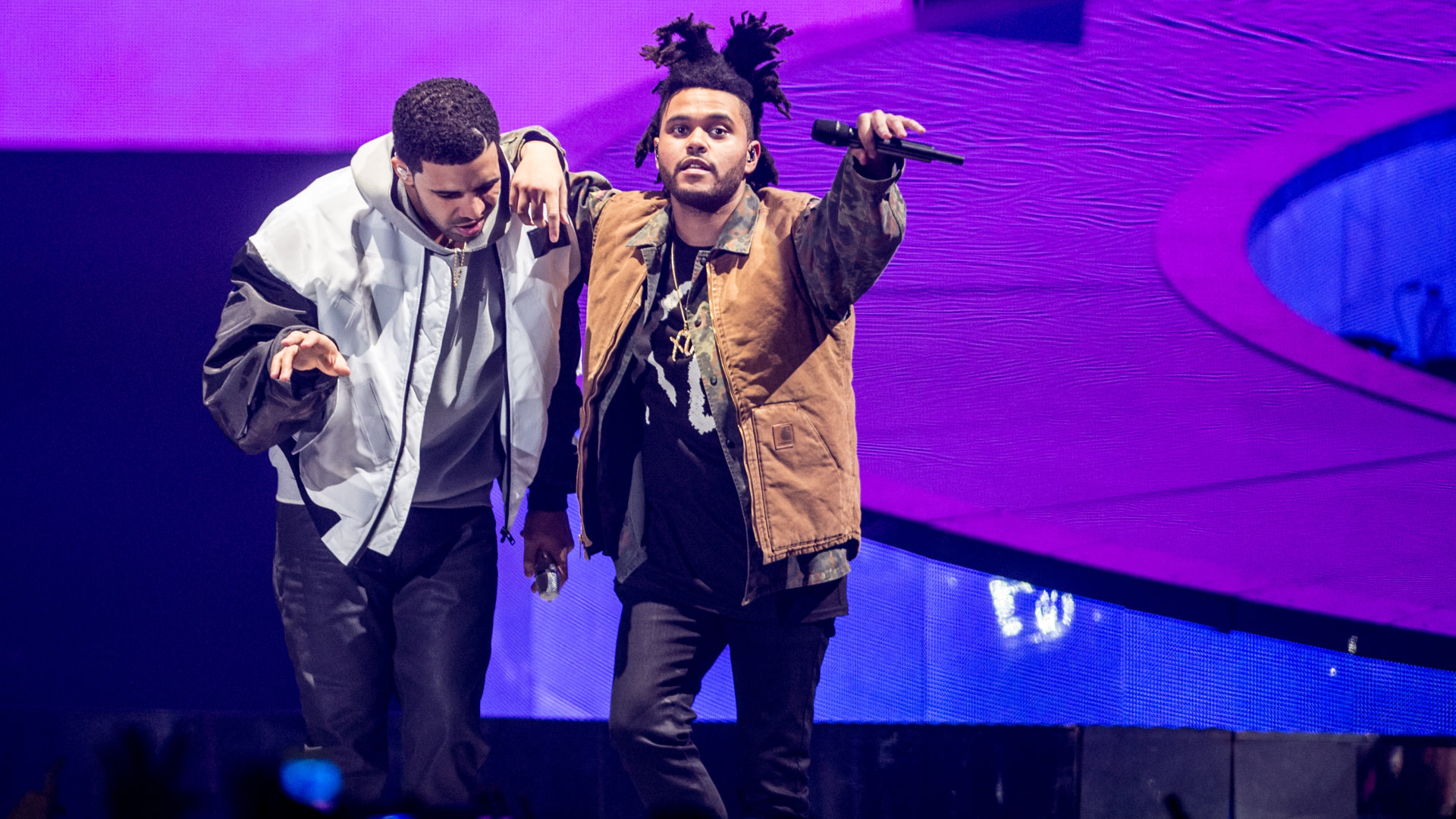 Drake and The Weeknd performing on stage together, with vibrant purple lighting in the background.