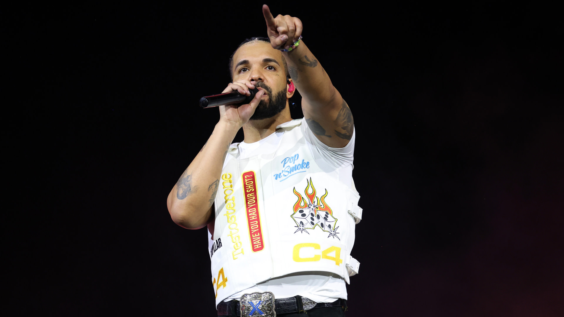 Drake performing on stage, wearing a white vest with graphics, holding a microphone, and pointing towards the audience.