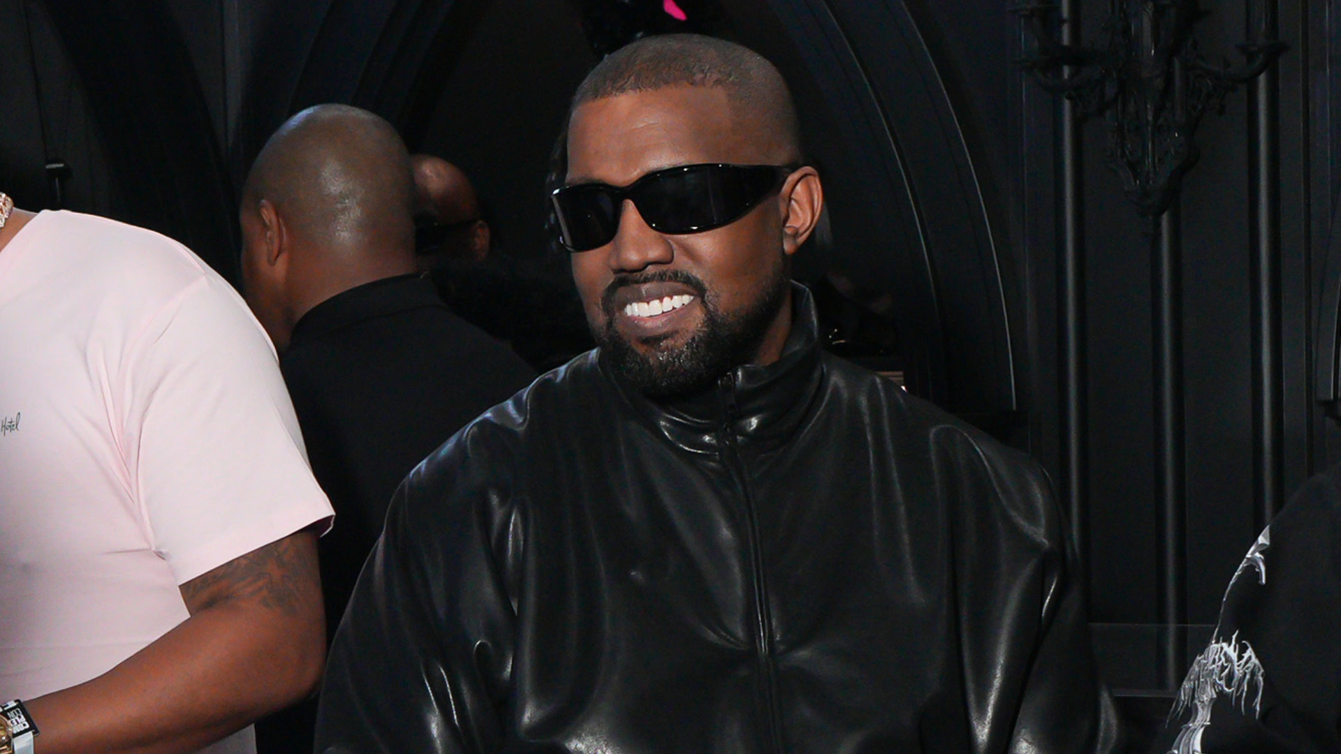 Ye wearing sunglasses and a black leather jacket, smiling indoors at an event.