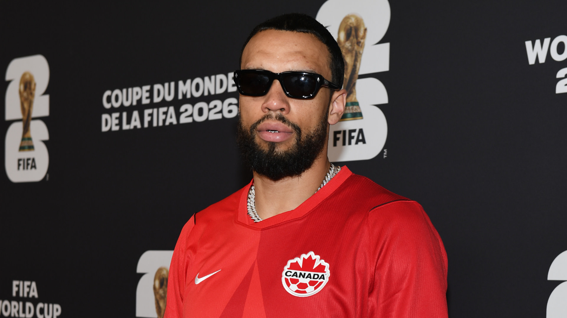 Dillon Brooks wearing sunglasses and a red Canada soccer jersey stands in front of a FIFA World Cup 2026 backdrop.