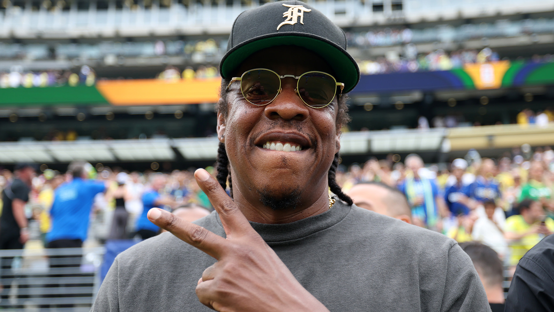 Jay-Z, wearing sunglasses and a cap, flashes a peace sign at a stadium event.