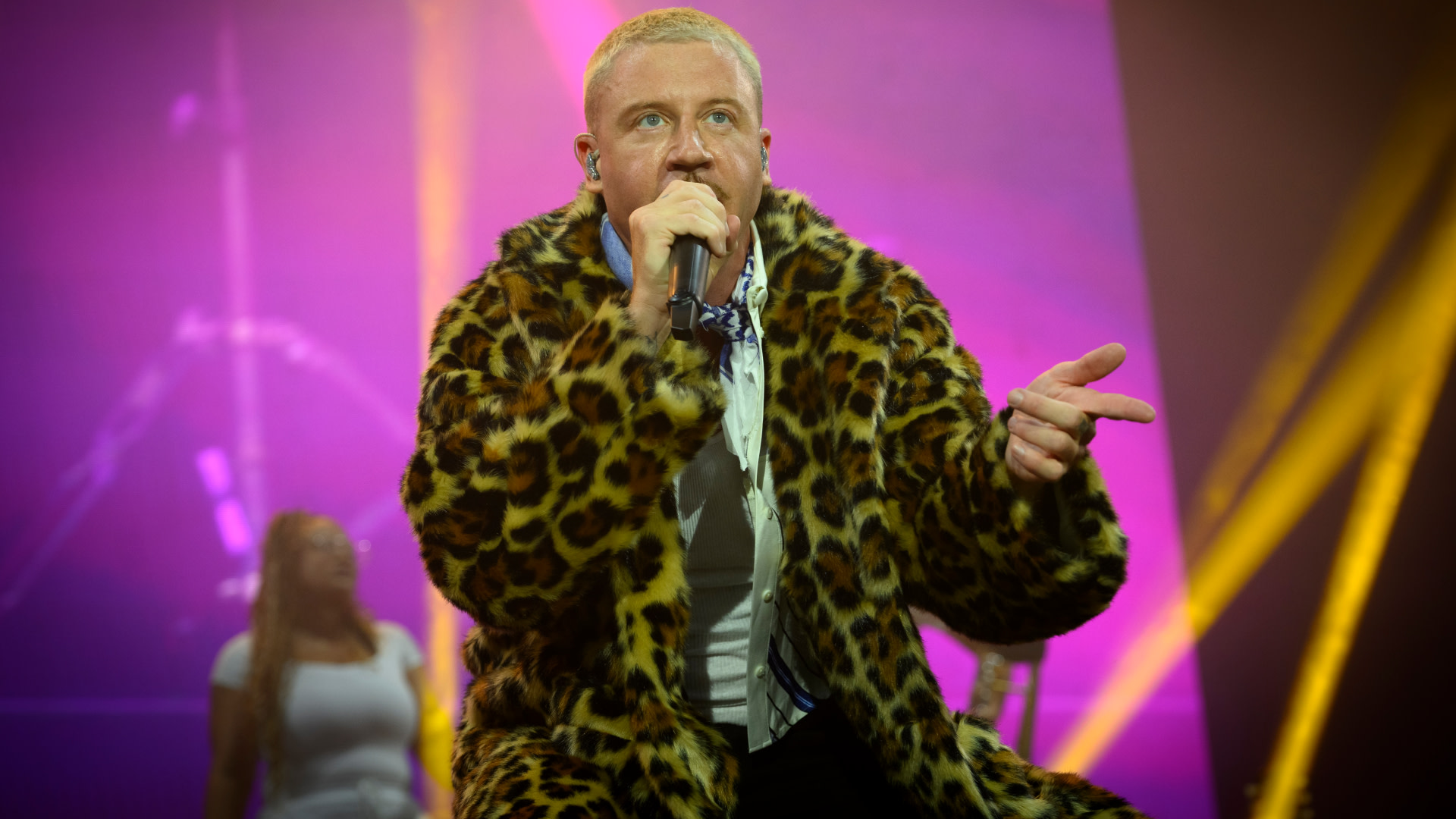 Macklemore performing on stage in a leopard print coat, holding a microphone, with colorful lights in the background.