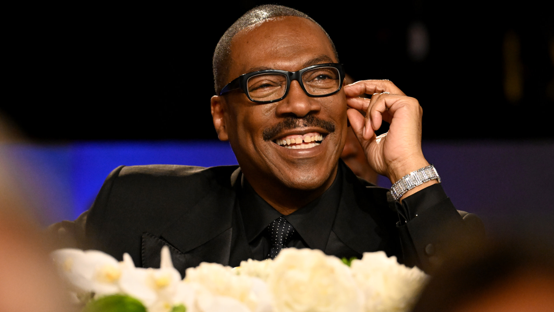 Eddie Murphy smiling, wearing glasses and a black suit, sitting at a table with white flowers.