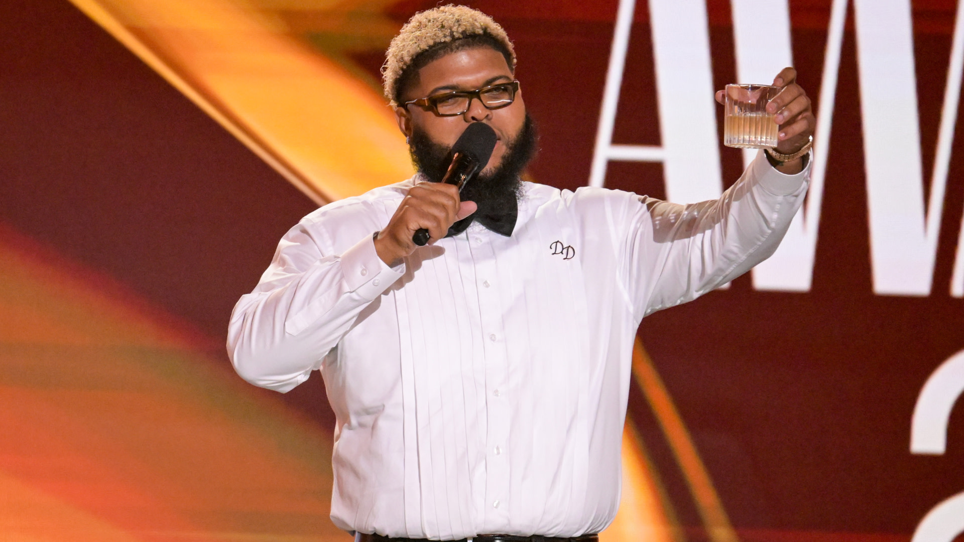 Druski with glasses and a beard, wearing a white shirt with "DD" on it, holds a microphone and a drink on stage at an awards event.
