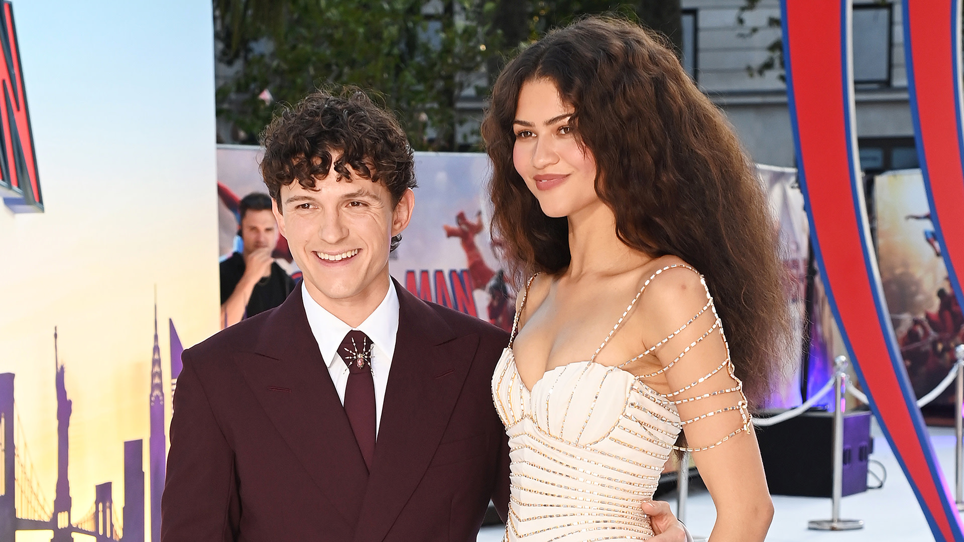 Tom Holland and Zendaya at a red carpet event. Tom wears a burgundy suit, and Zendaya is in a white dress with pearl straps.
