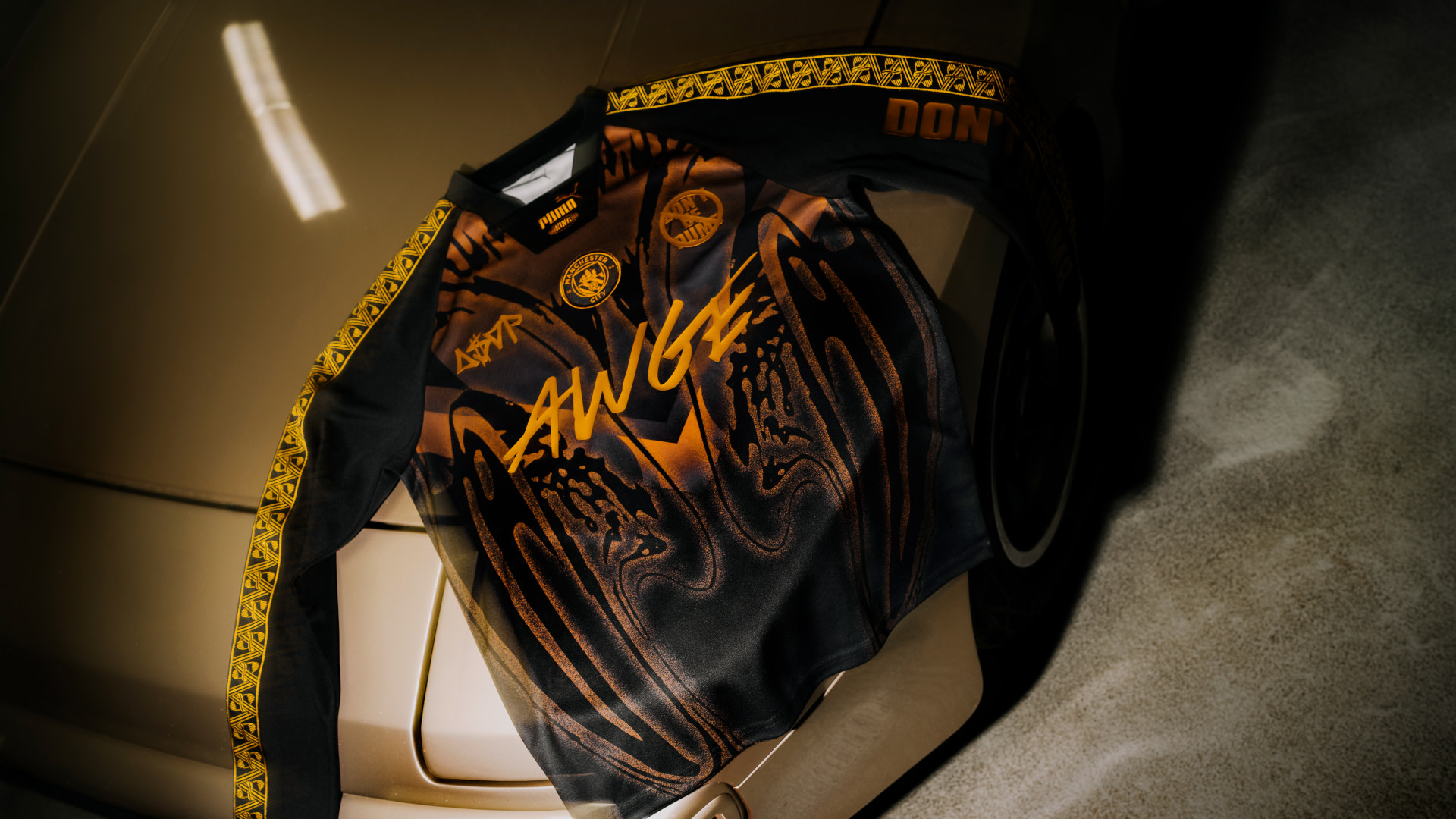 A stylish black and orange long-sleeve shirt with "AWGE" and various logos, draped over a beige car hood in a dimly lit setting.