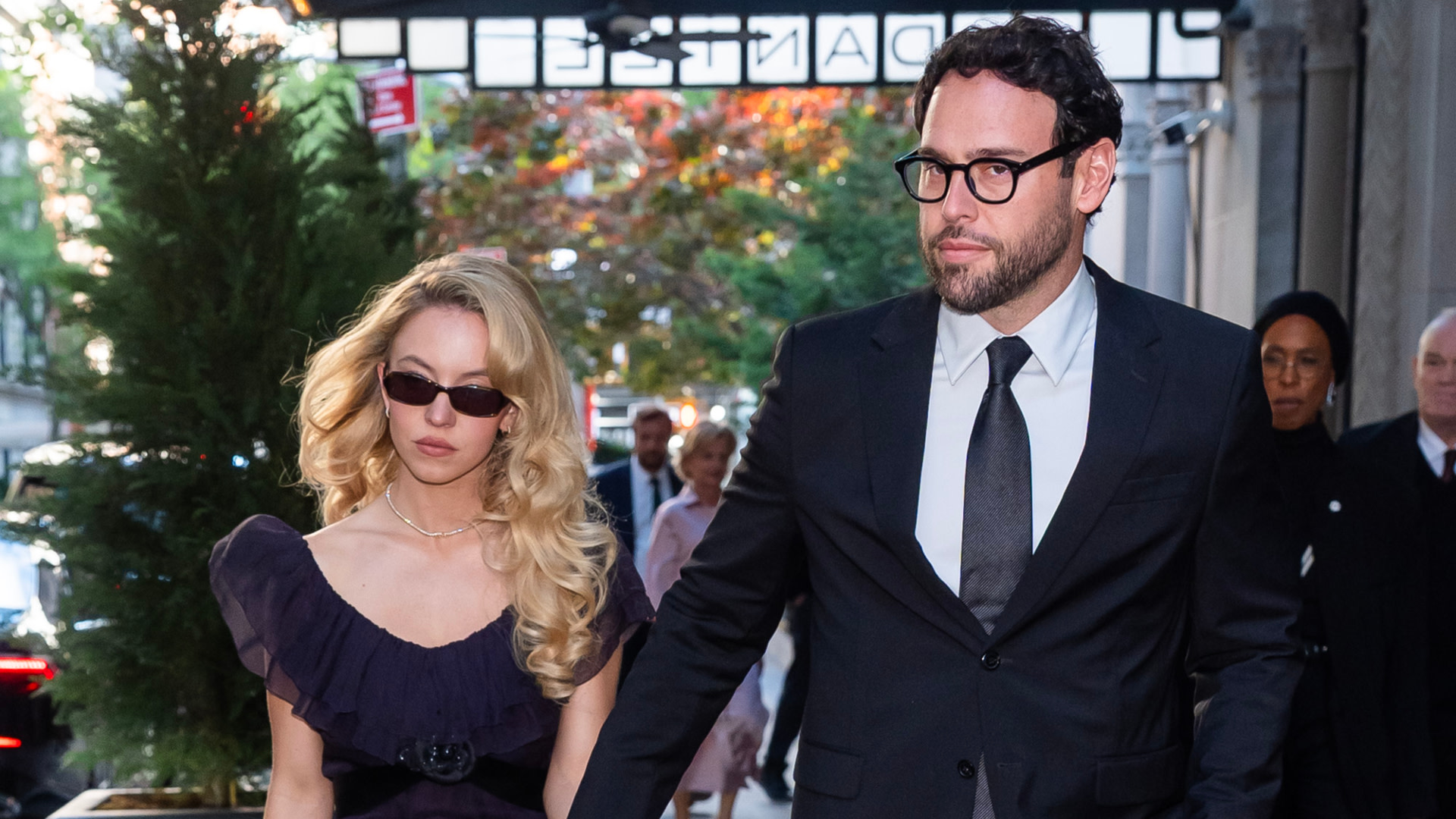 Scooter Braun in a suit and Sydney Sweeney in a black dress and sunglasses walking together, with a tree and people in the background.