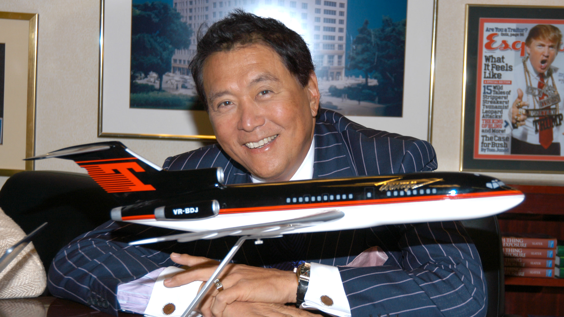 Robert Kiyosaki in a suit smiles beside a model airplane, with framed pictures and magazines in the background.