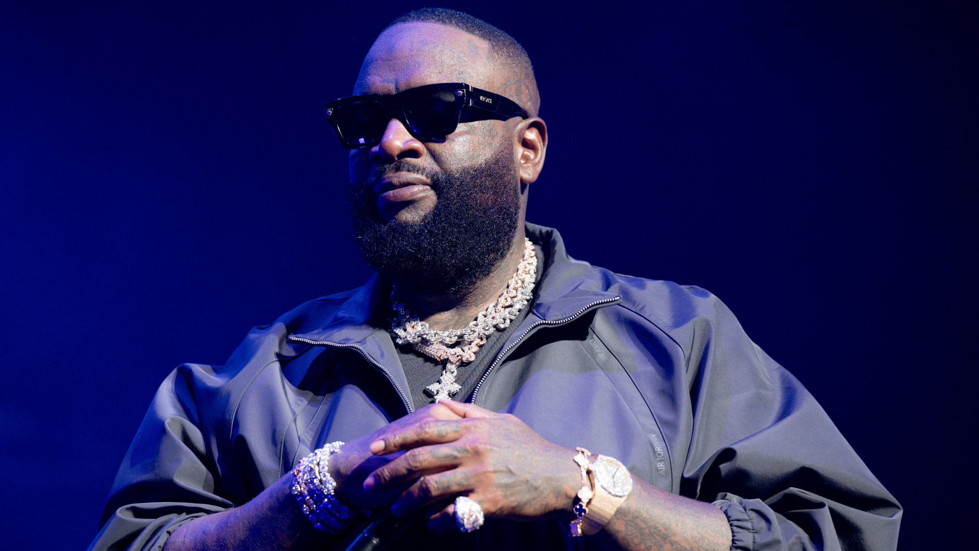 Rapper Rick Ross on stage wearing sunglasses, a dark jacket, and multiple necklaces, with a confident expression.