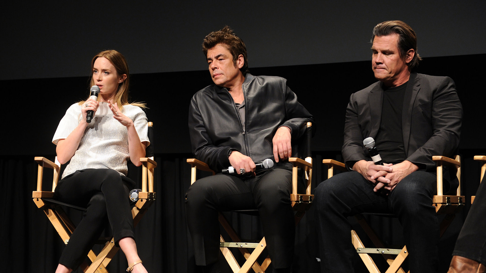 Emily Blunt, Benicio Del Toro, and Josh Brolin sit on stage in director chairs, each holding microphones.