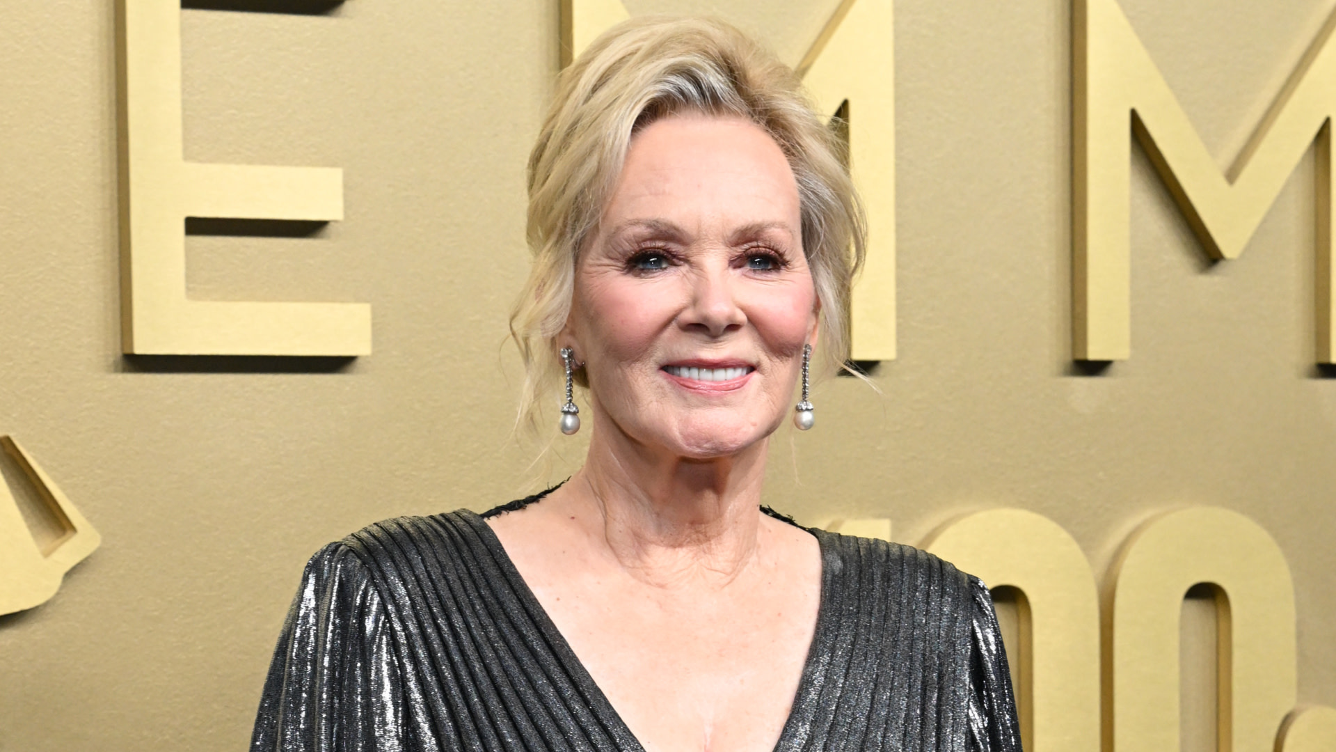 Jean Smart in a metallic dress, smiling at an event with an "EMMY" backdrop.
