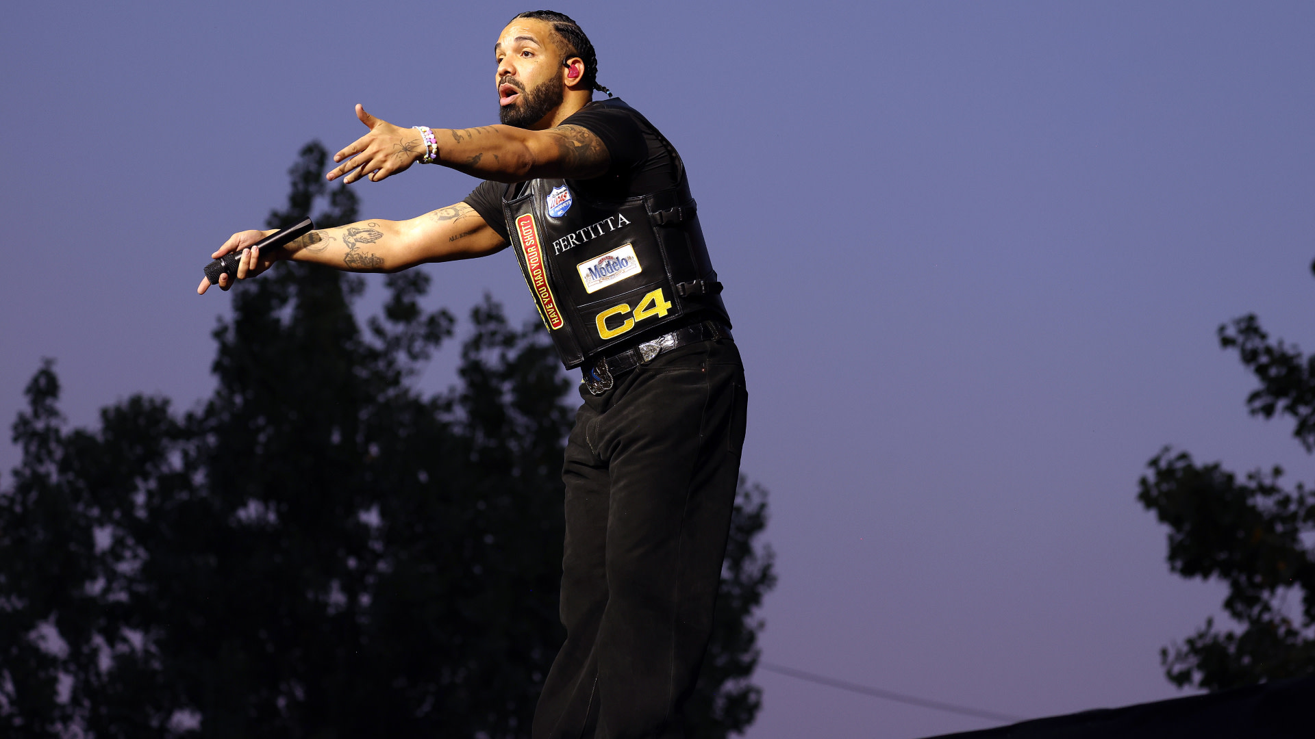Drake on stage wearing a black vest with patches, gesturing with both hands. Trees and a clear sky are in the background.