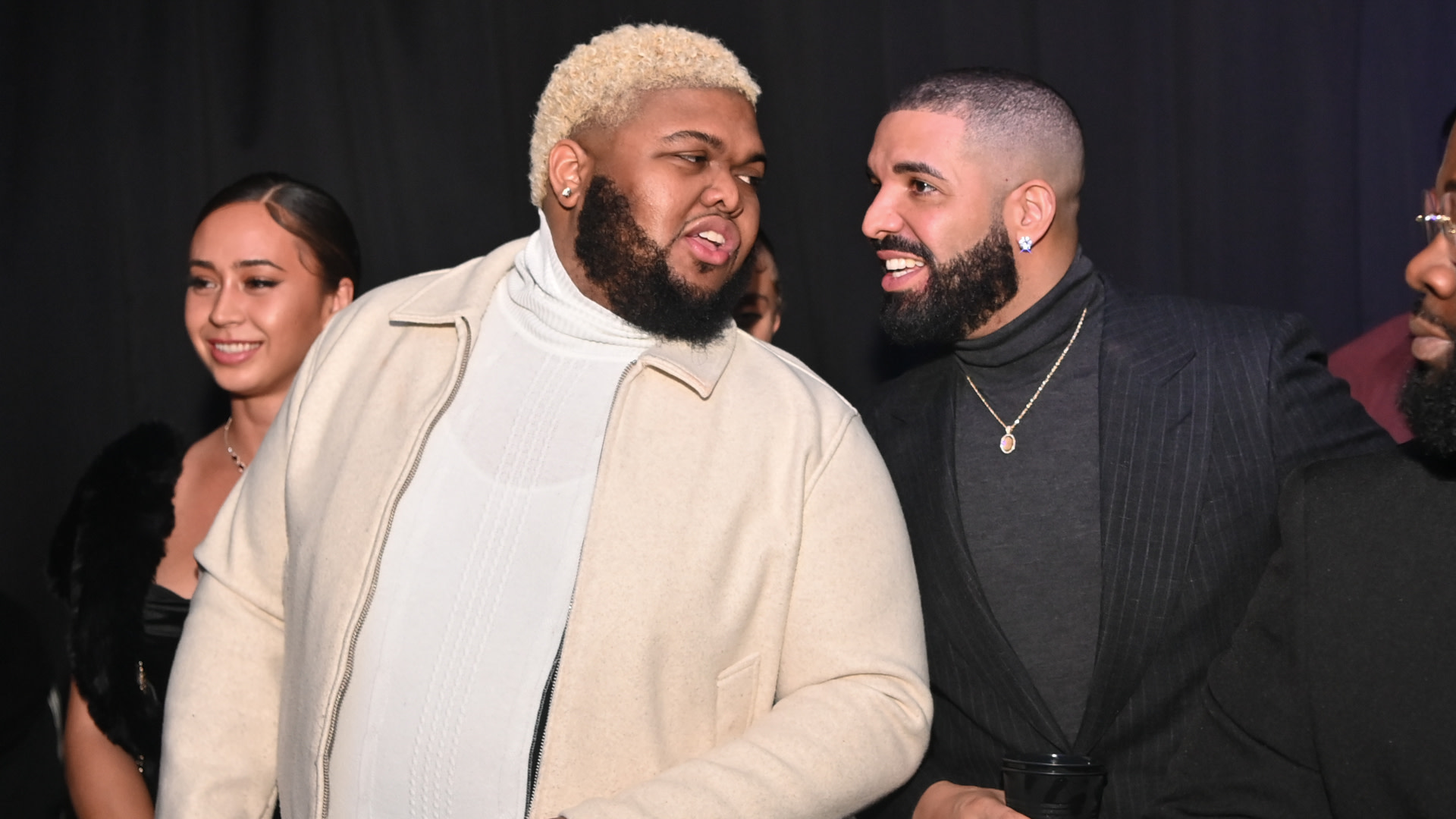 Druski and Drake are smiling and talking at an event, with a woman in the background.