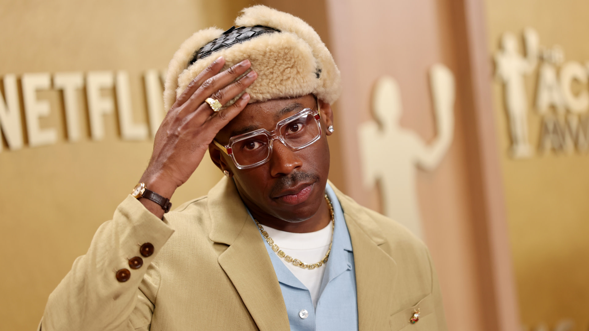 Tyler wearing a beige suit, fur hat, and clear glasses, posing at a Netflix event backdrop.