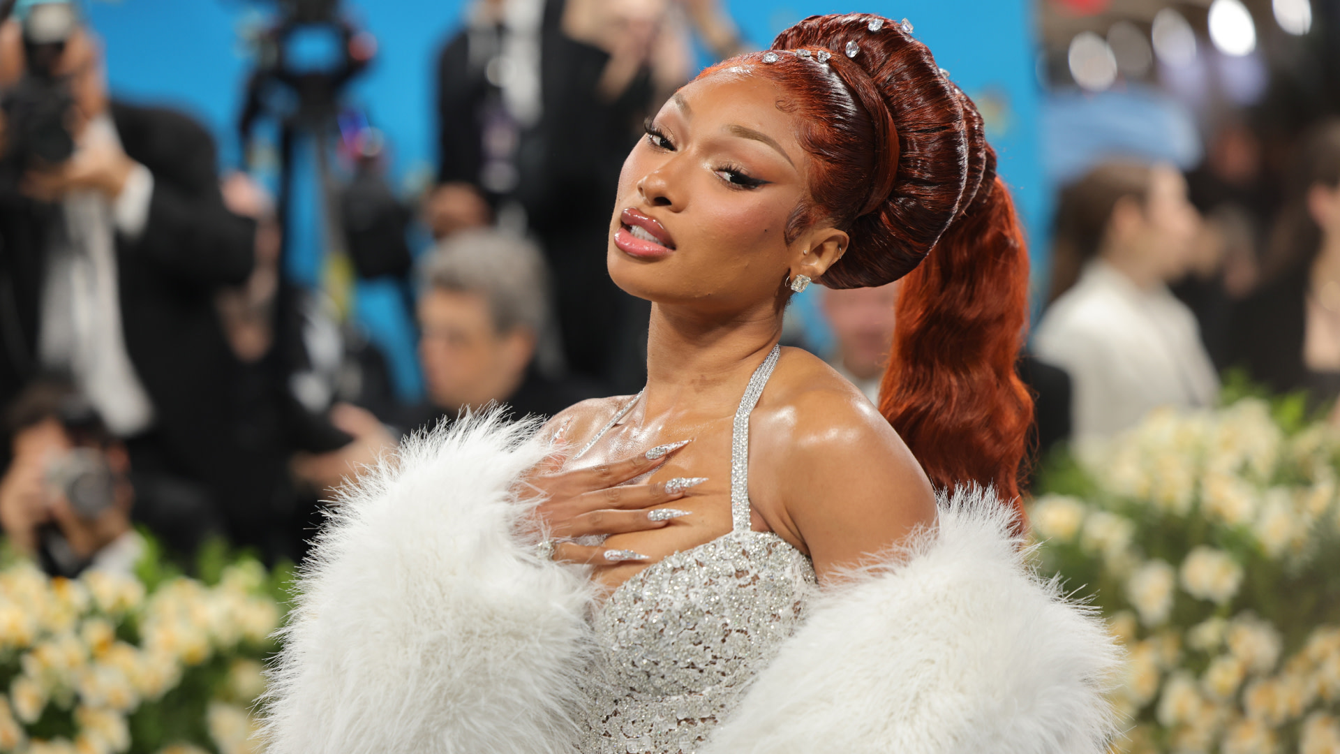 Megan Thee Stallion with red hair in an elegant gown and white fur wrap poses confidently at an event, with a blurred background of people and flowers.