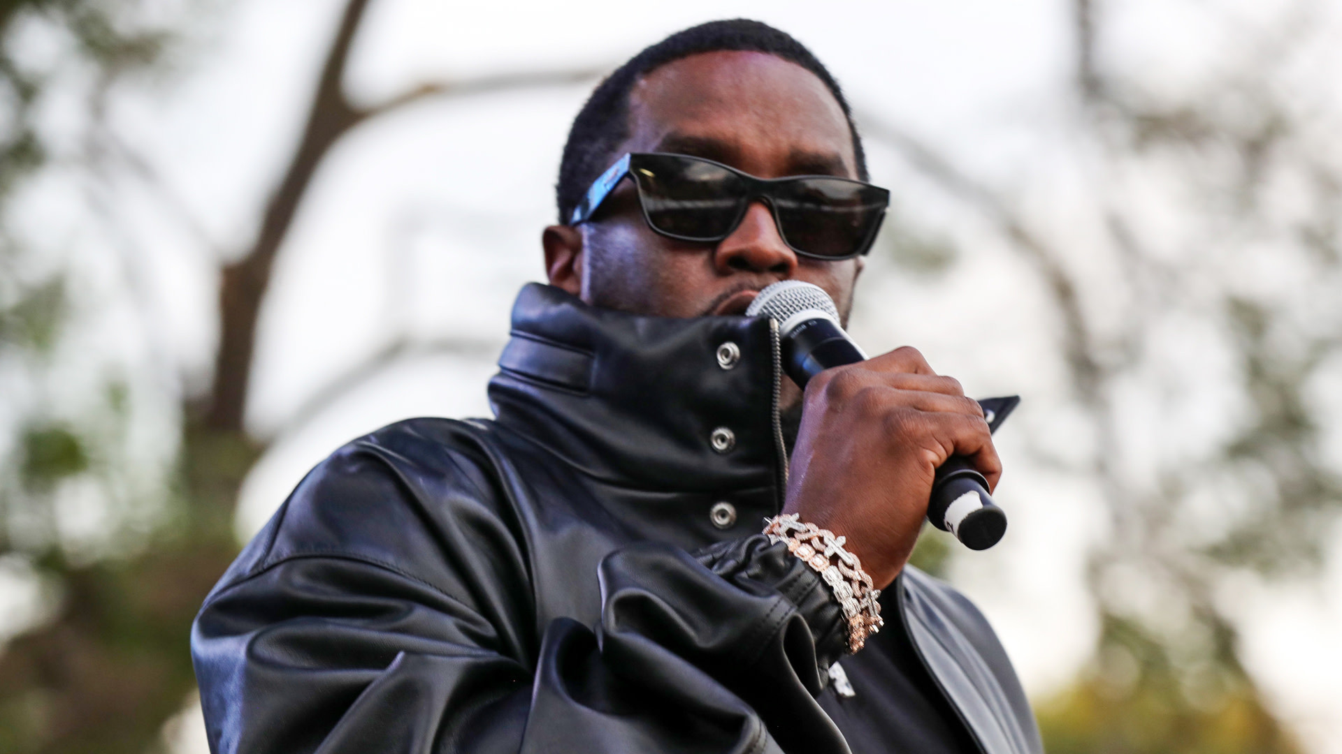 Diddy in a black leather jacket and sunglasses holds a microphone outdoors.