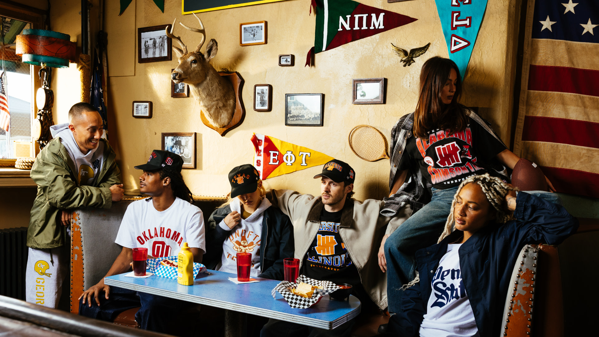 A group of people in casual, collegiate-style clothing sits in a diner booth with sports memorabilia and pennants on the walls.