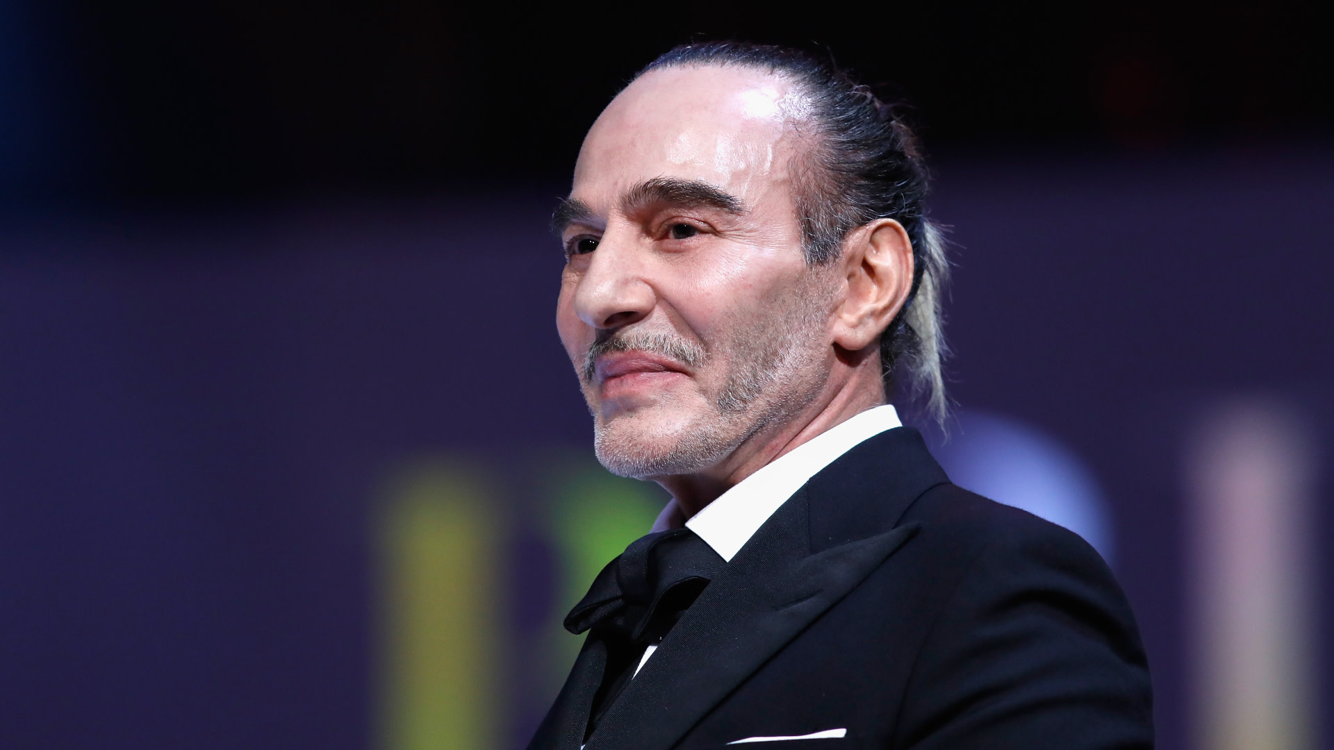 A man in a suit with a slicked-back hairstyle and a subtle smile, against a blurred background.