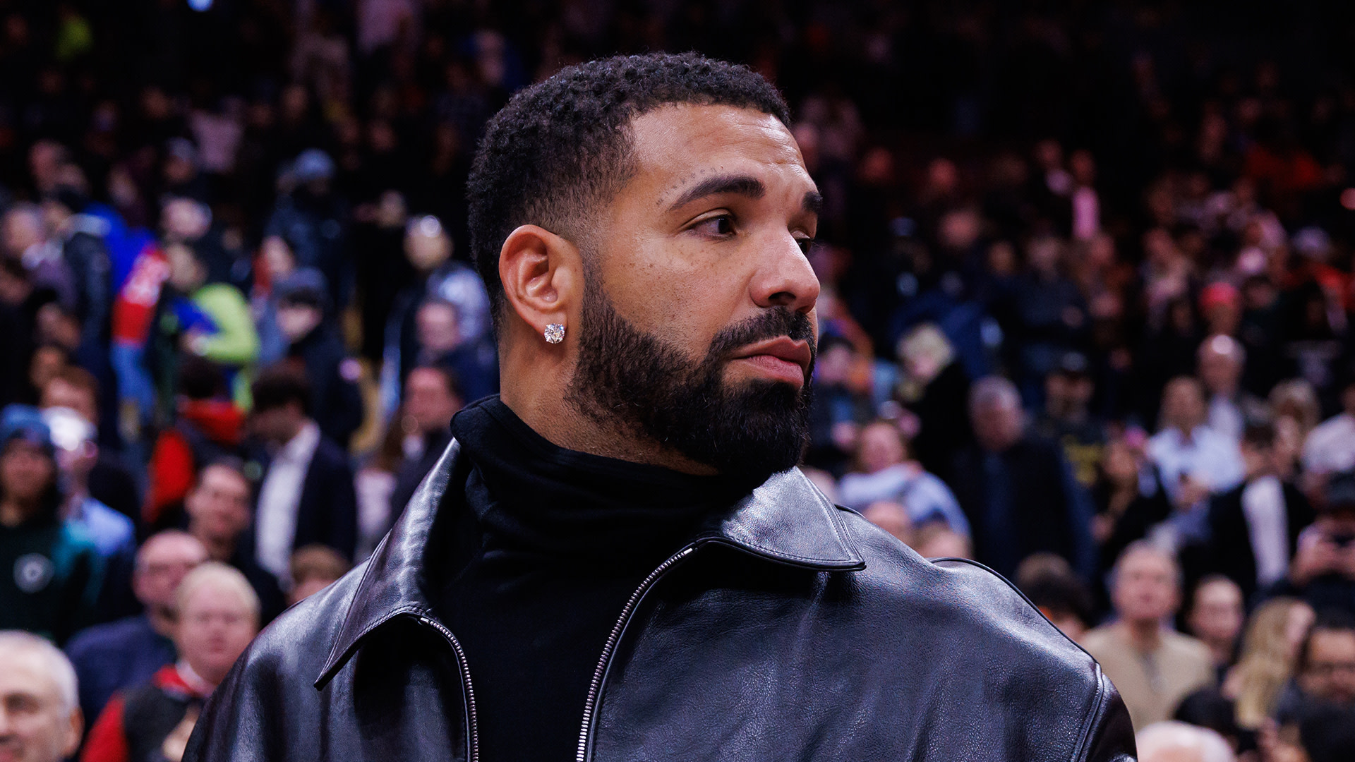 Drake in a black leather jacket, looking to the side, with a crowd in the background.