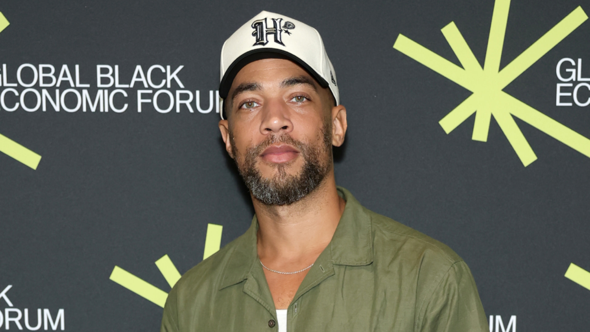 Kendrick Sampson wearing a white cap and green shirt stands in front of a backdrop with "Global Black Economic Forum" text.