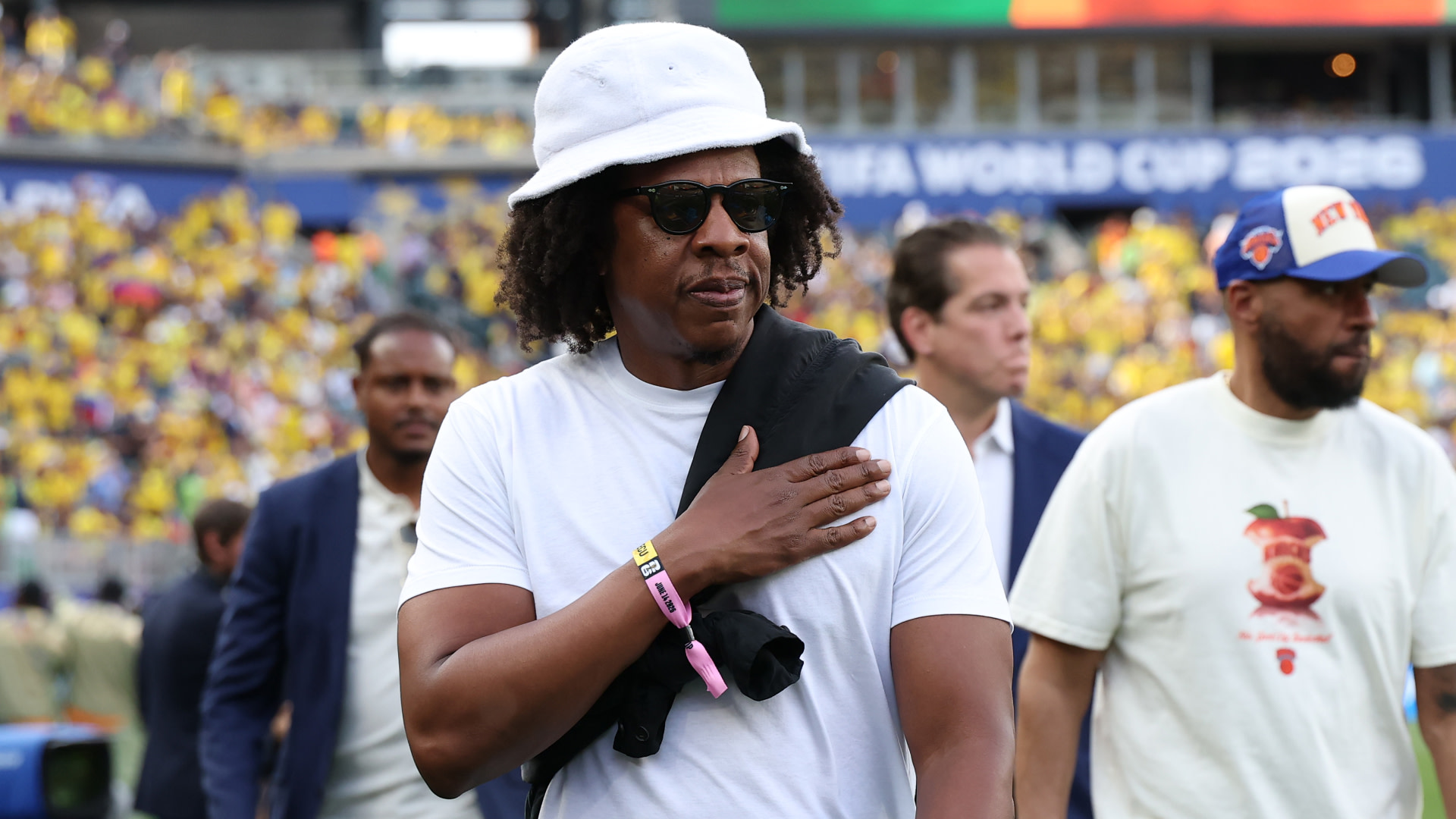 Jay-Z wearing a white bucket hat and sunglasses at a sports event, with a crowd in the background.