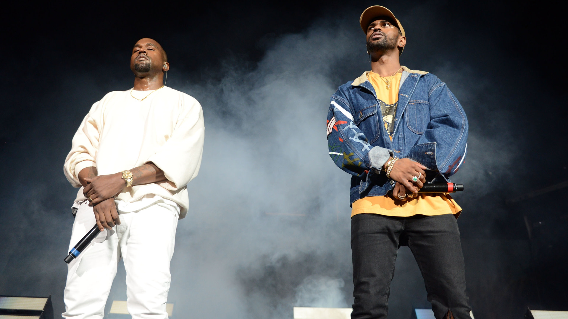 Kanye West and Big Sean performing on stage, surrounded by smoke, holding microphones, wearing casual outfits.