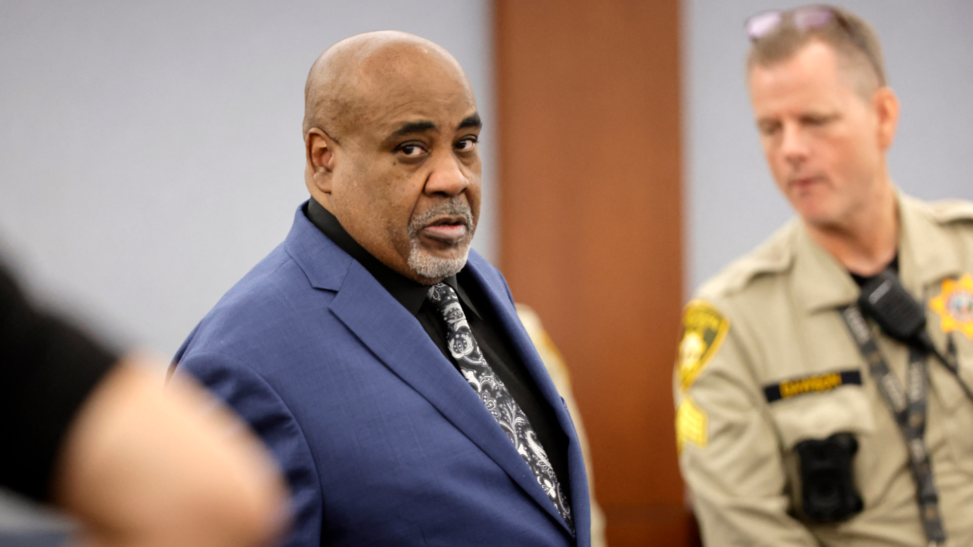 Keffe D in a blue suit stands in a courtroom, with a law enforcement officer in uniform nearby.