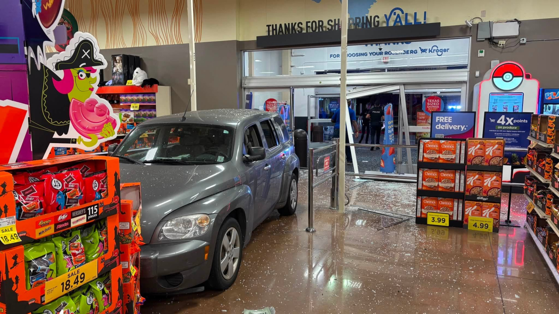 A car has crashed into a grocery store entrance, surrounded by Halloween-themed displays and snack boxes.