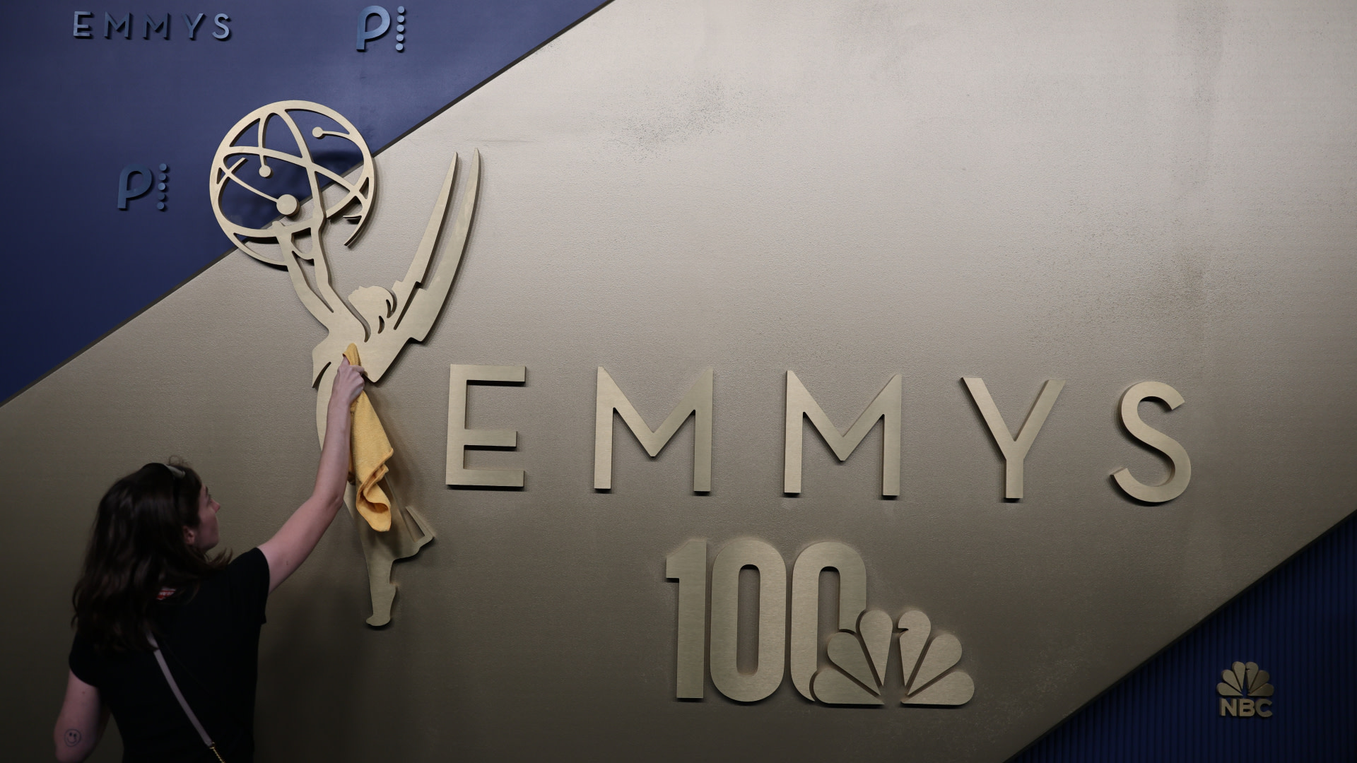A person cleaning an Emmy Awards logo on a wall, with "EMMYS" and "100" prominently displayed. NBC logo visible.