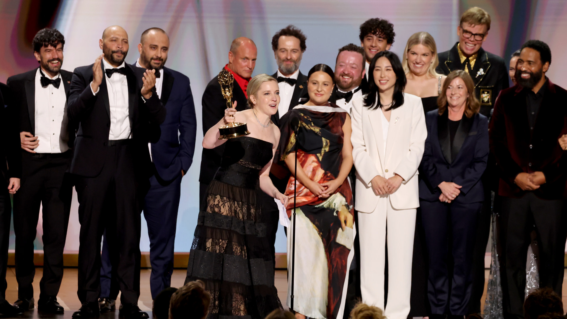 A group of people on stage at an awards ceremony, with one woman holding an Emmy. They are dressed in formal attire, smiling and celebrating.