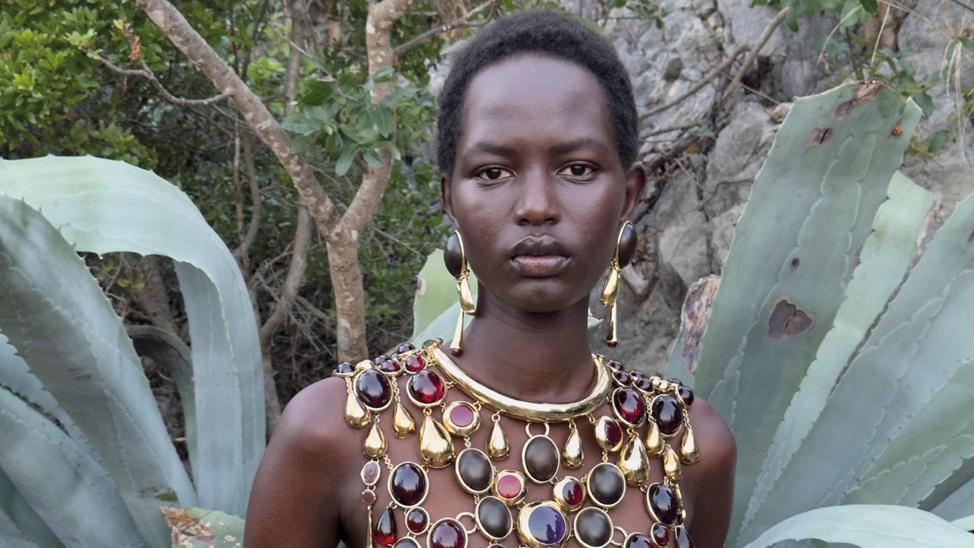 A woman stands in front of large agave plants, wearing a dress adorned with large gemstones and gold accents.