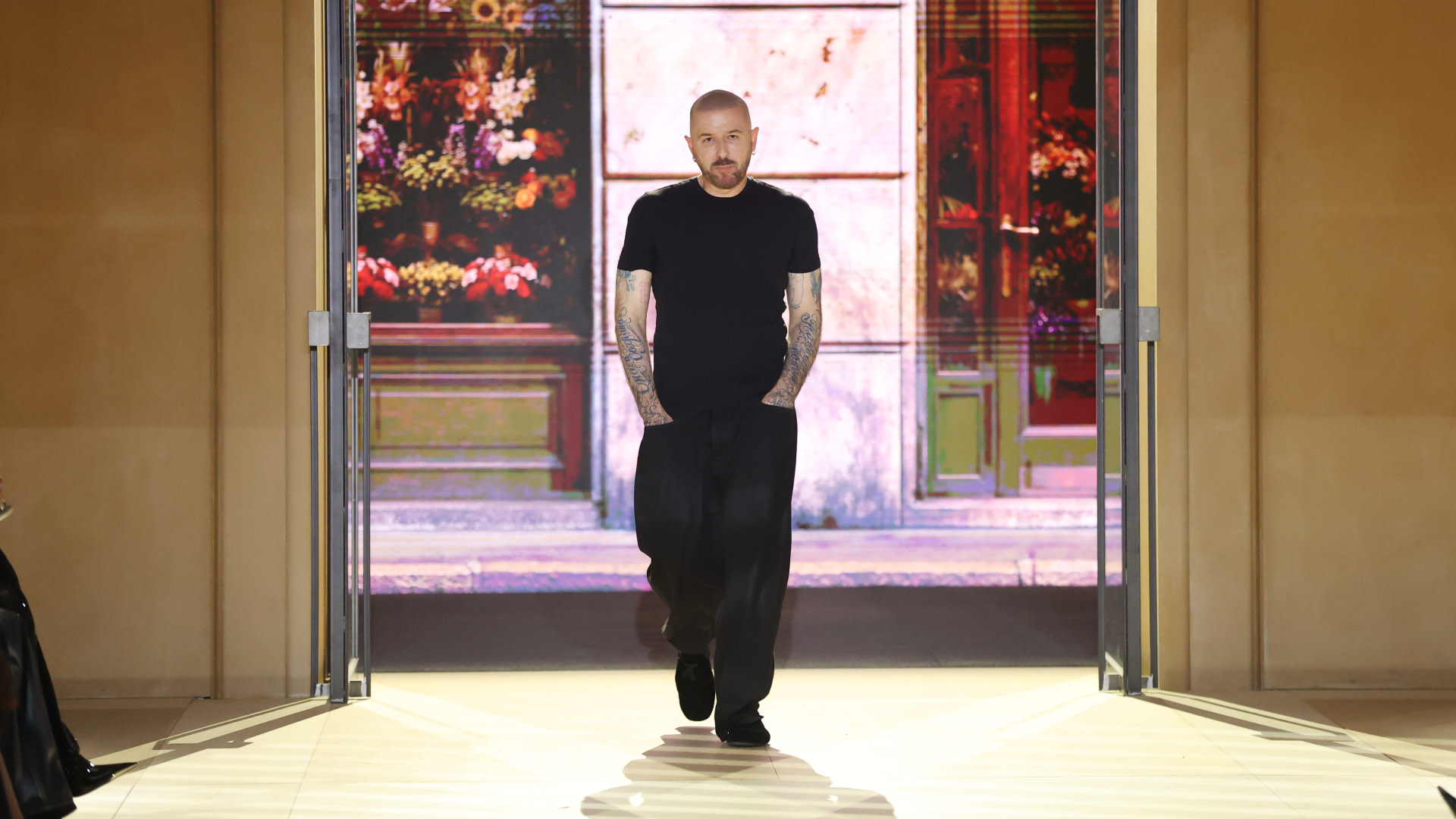 Demna with tattoos and a black outfit walks on a runway with a floral backdrop.