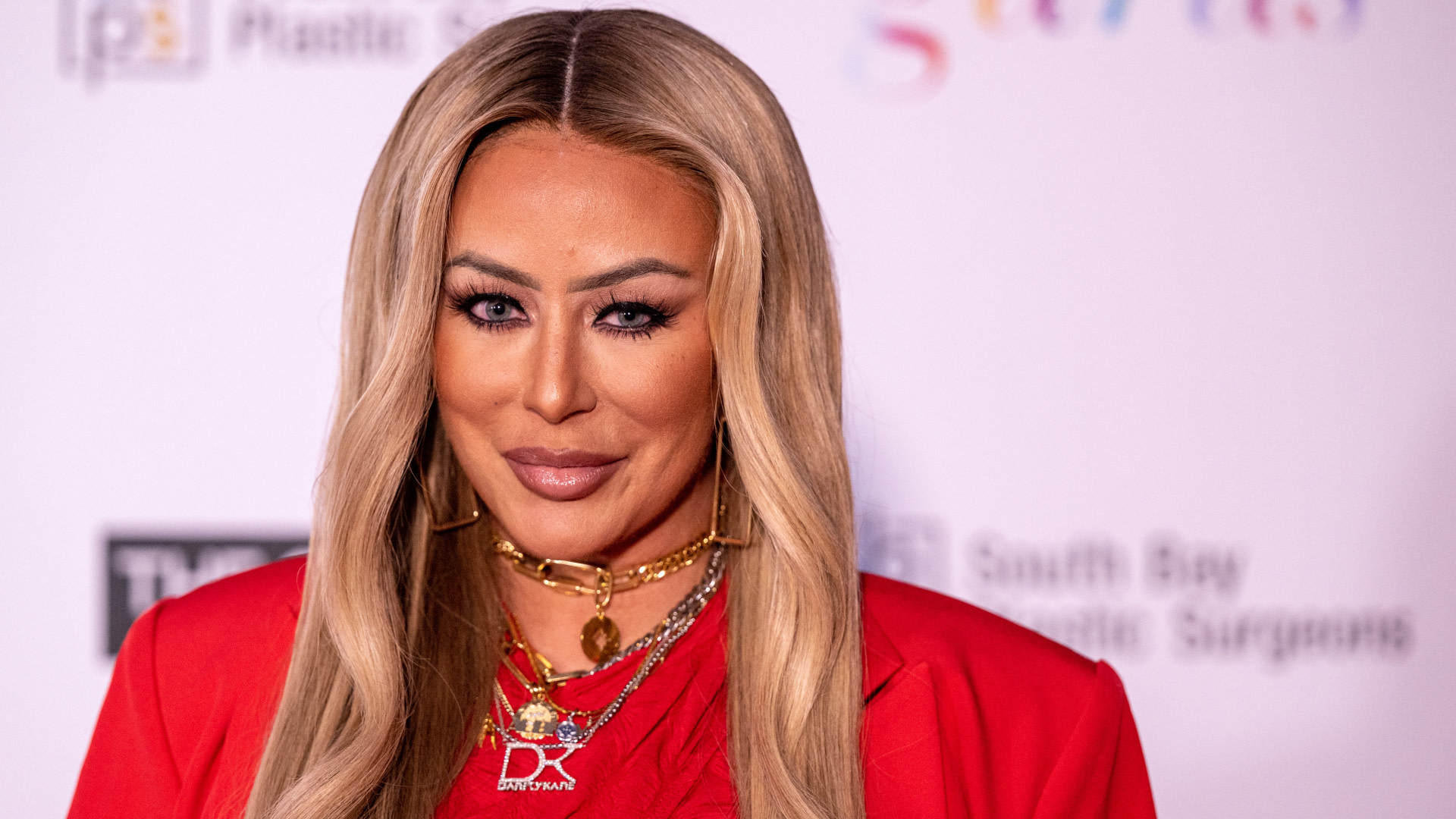 Aubrey O'Day with long blonde hair wearing a red outfit and layered gold necklaces at an event.