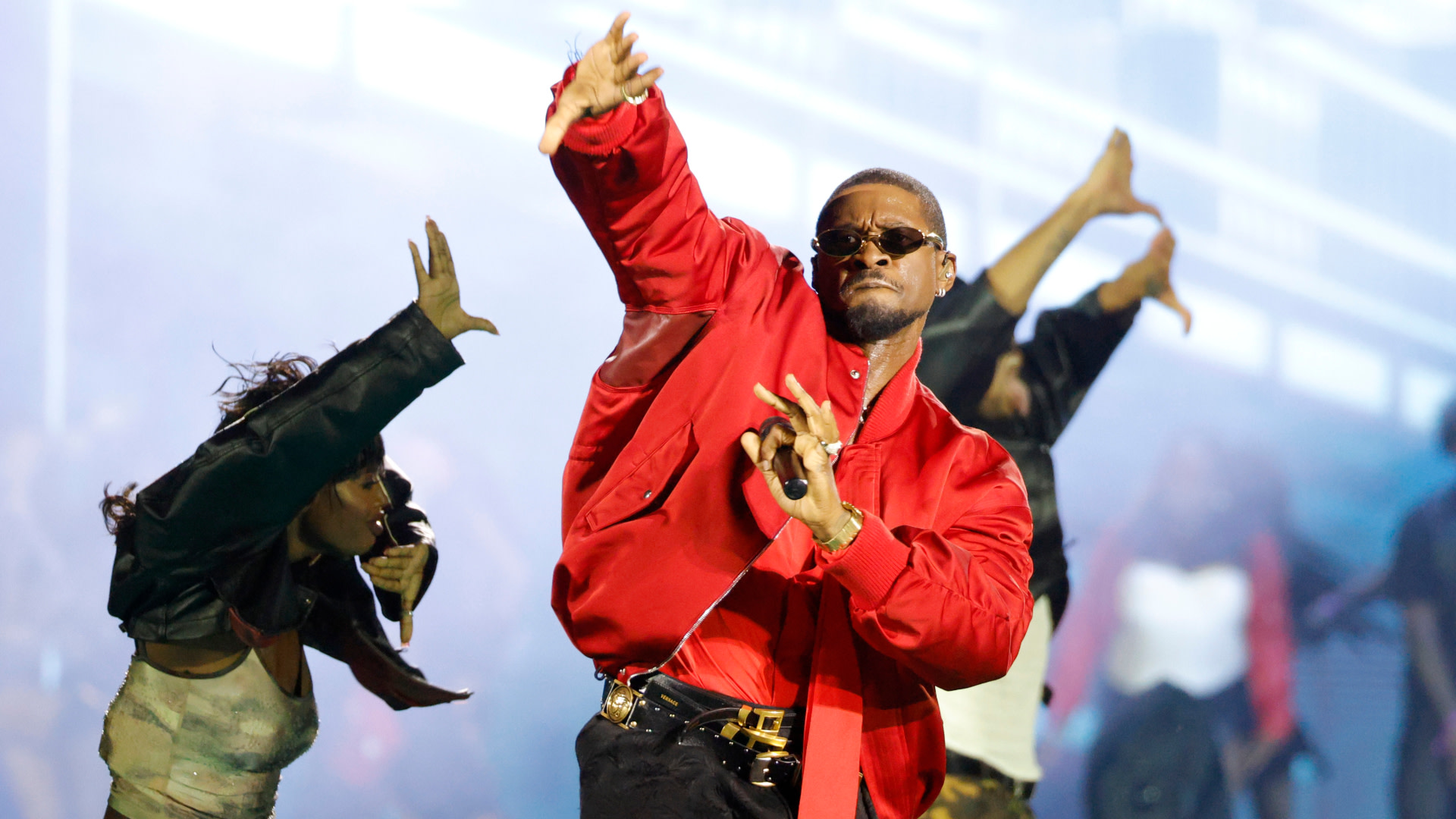 Usher performs energetically on stage in a red jacket and sunglasses, surrounded by dancers.