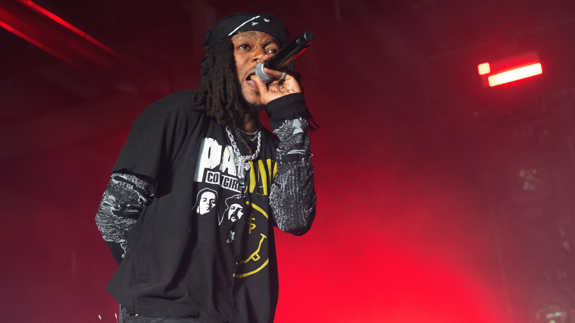JID with dreadlocks and a black shirt sings into a microphone on stage with red lighting.