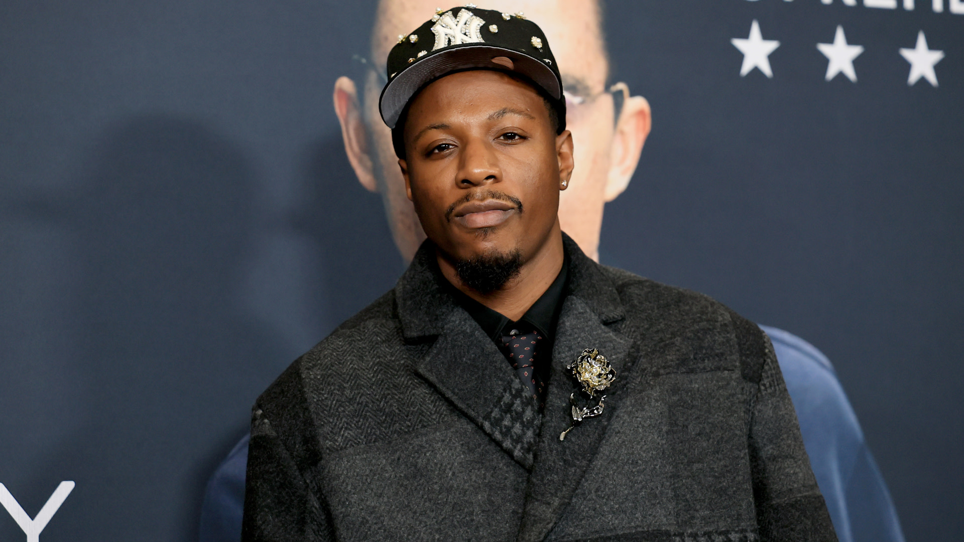 Joey Badass wearing a dark coat and a patterned cap at an event, standing against a backdrop with stars.