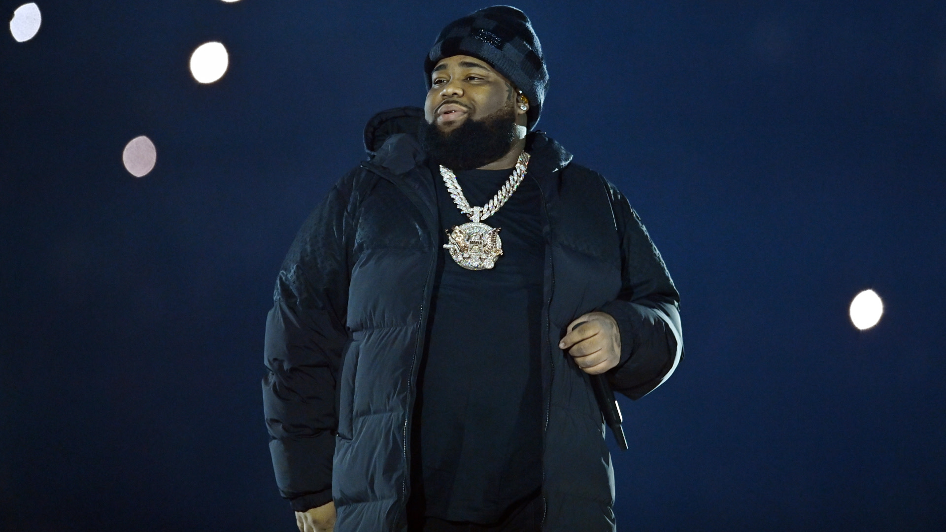 Rod Wave in a black jacket and beanie stands on stage with a large chain necklace, against a dark background with bright lights.