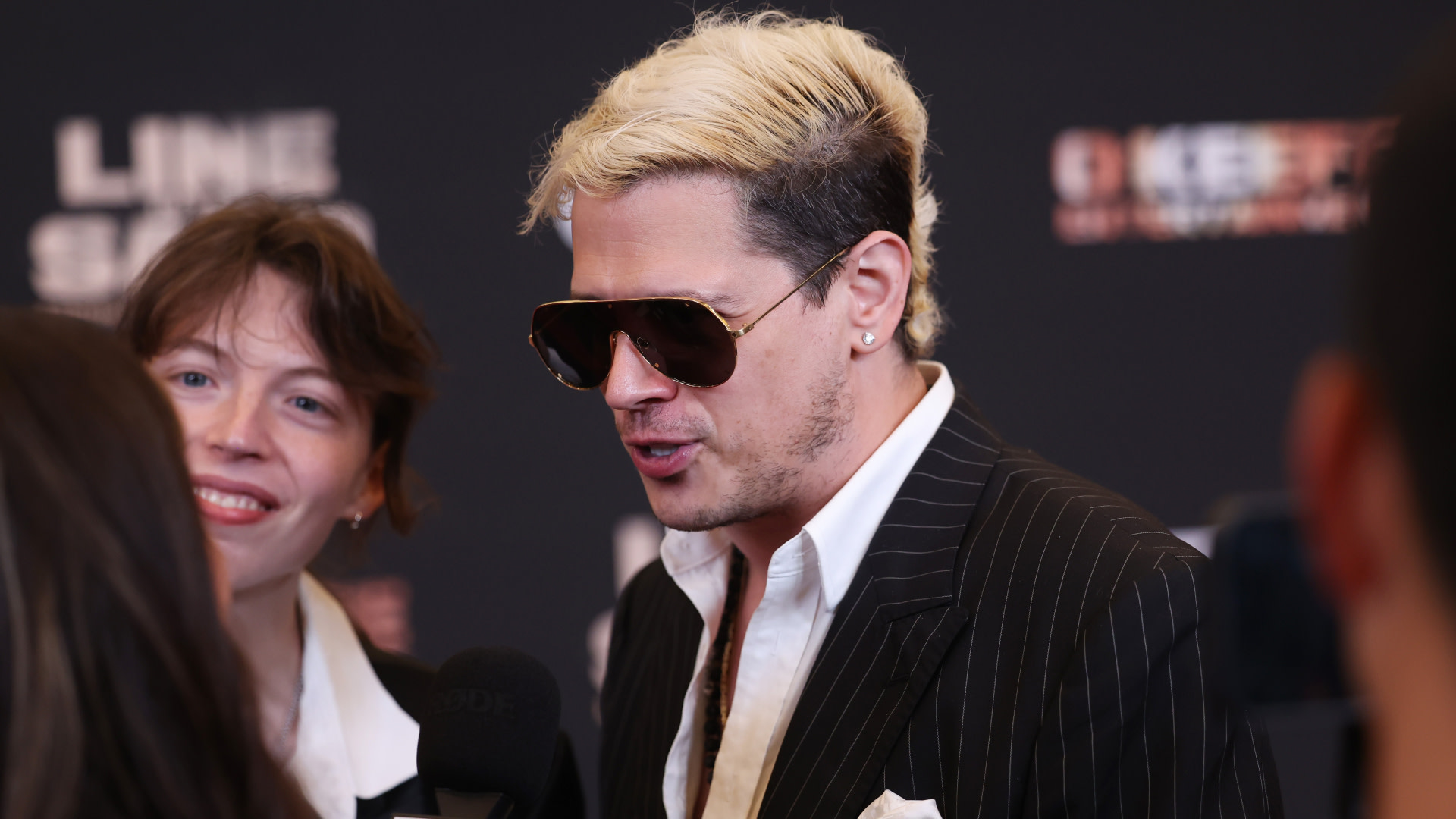 Milo with blond hair and sunglasses in a pinstripe suit speaks at an event, with another person smiling beside them.
