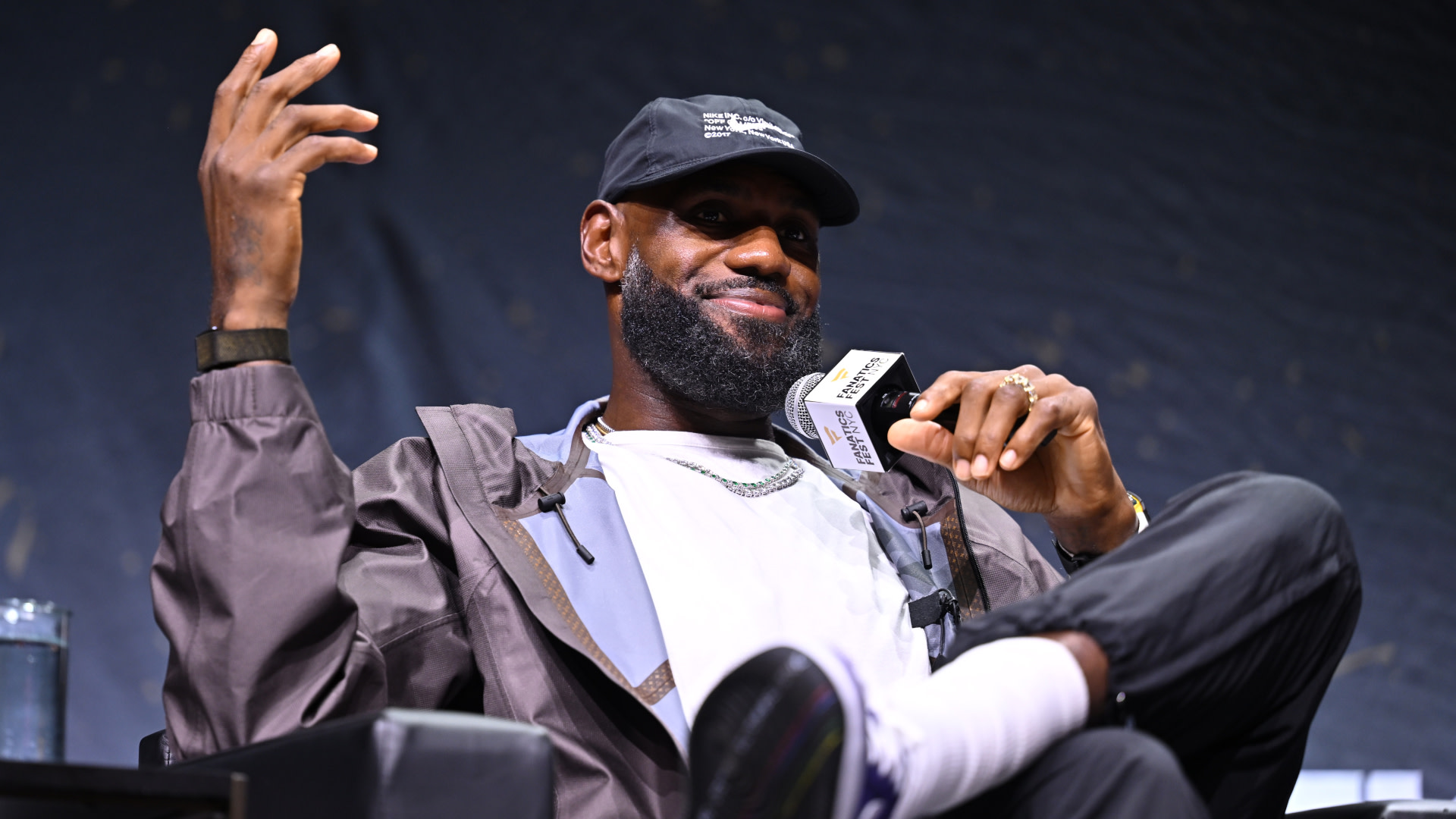 LeBron James, wearing a cap and casual jacket, speaks at an event, holding a microphone with a relaxed expression.