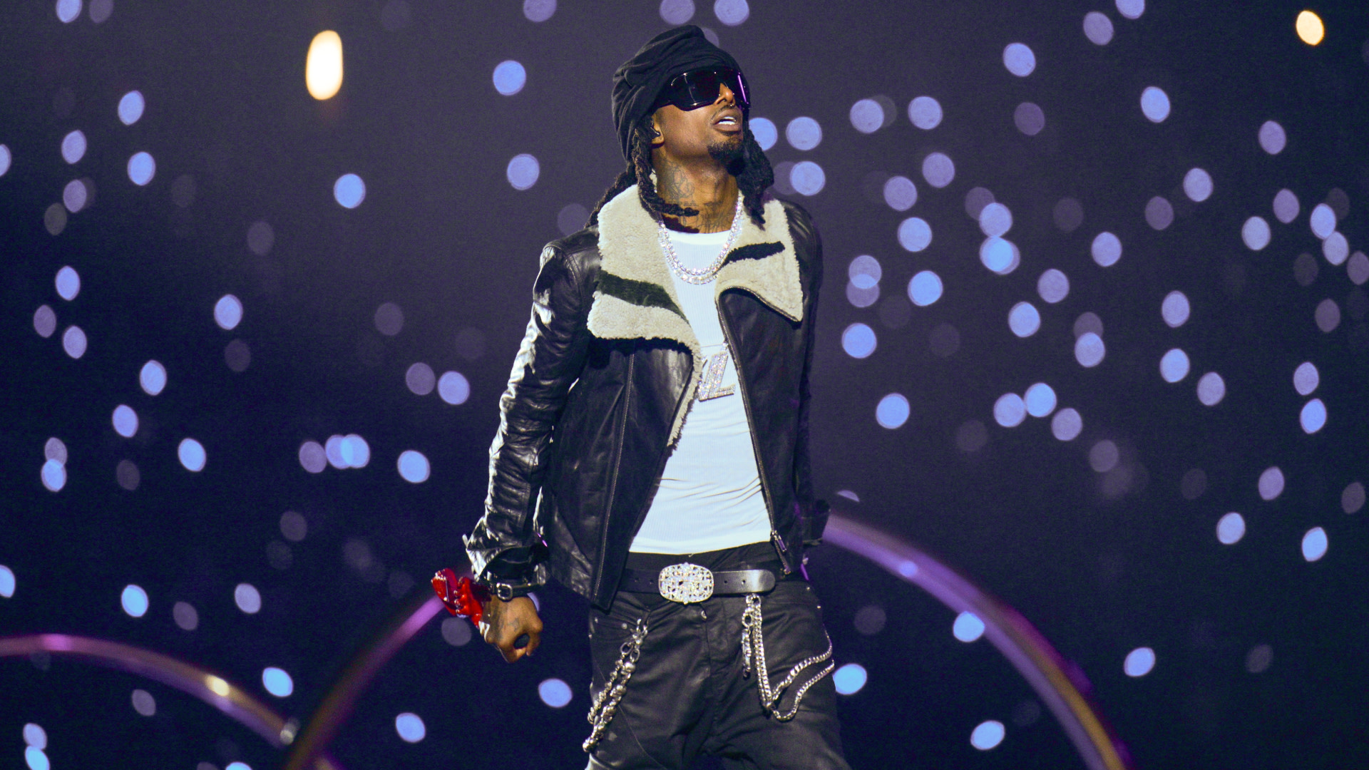 Playboi Carti on stage wearing a black leather jacket and sunglasses, with a backdrop of sparkling lights.