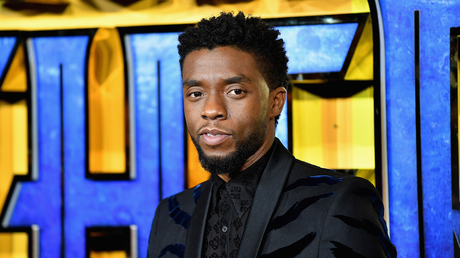 Chadwick Boseman at an event, wearing a dark suit with a patterned shirt, standing in front of a colorful background.