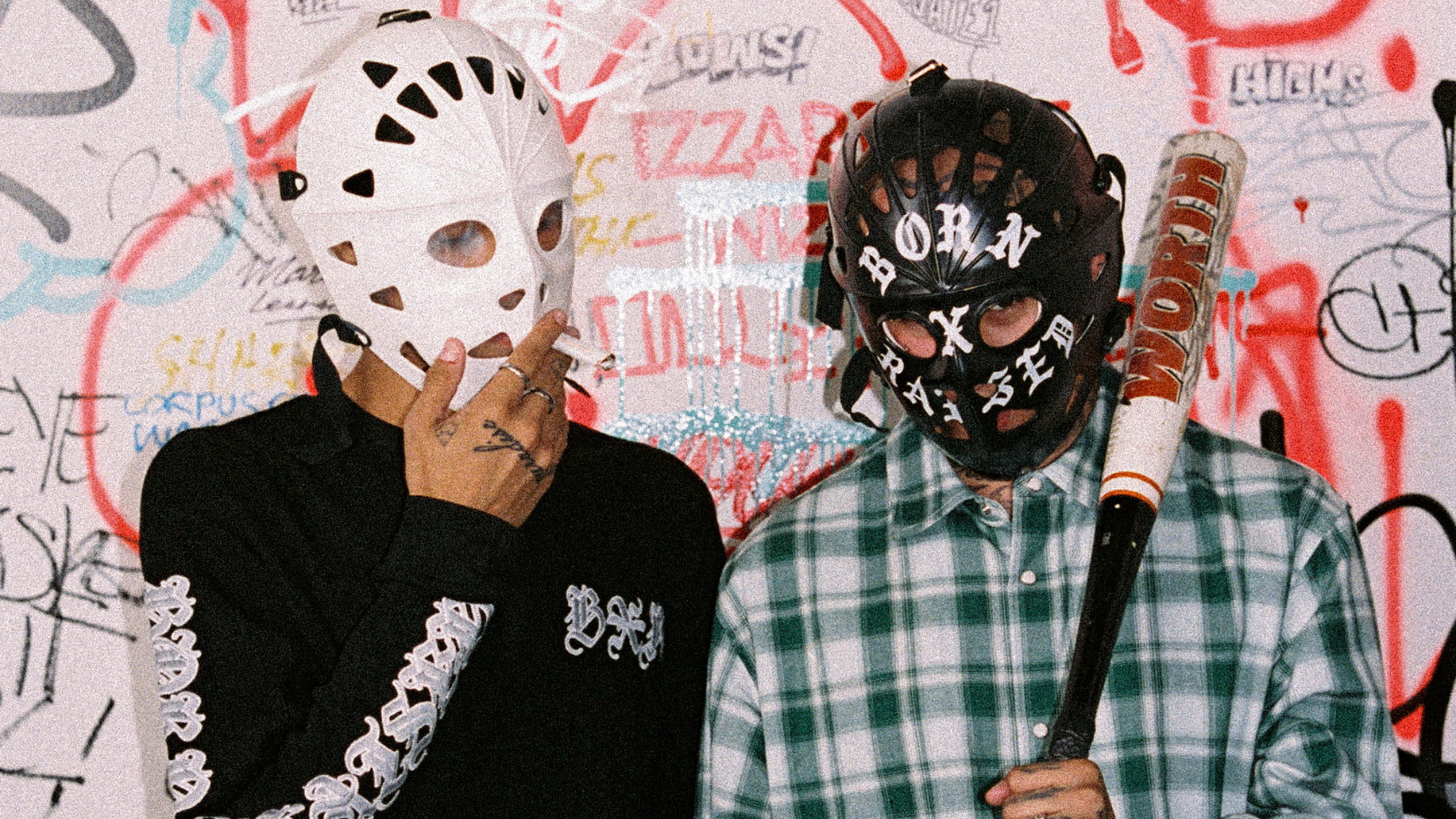 Two people in hockey masks, one smoking and the other holding a bat, stand in front of a graffiti-covered wall.