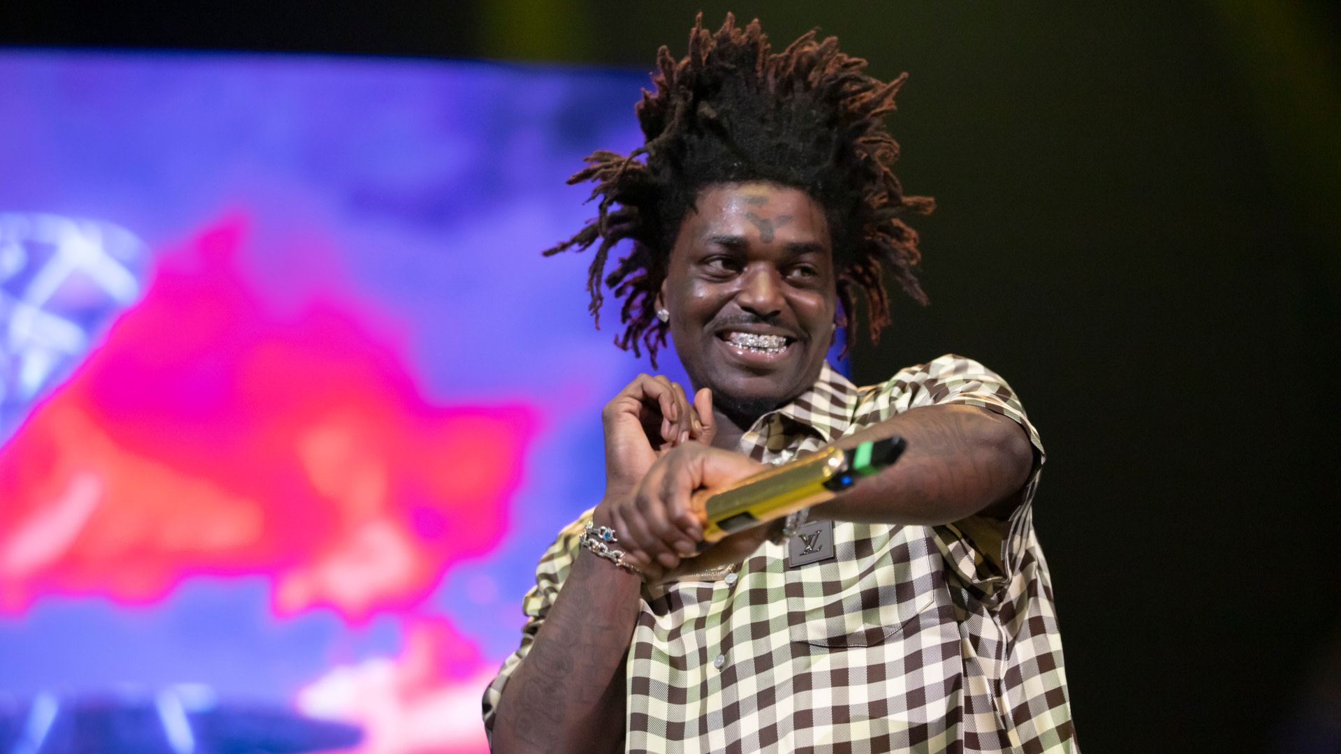 Kodak Black with spiky hair and a checkered shirt smiles on stage, holding a microphone with colorful lights in the background.