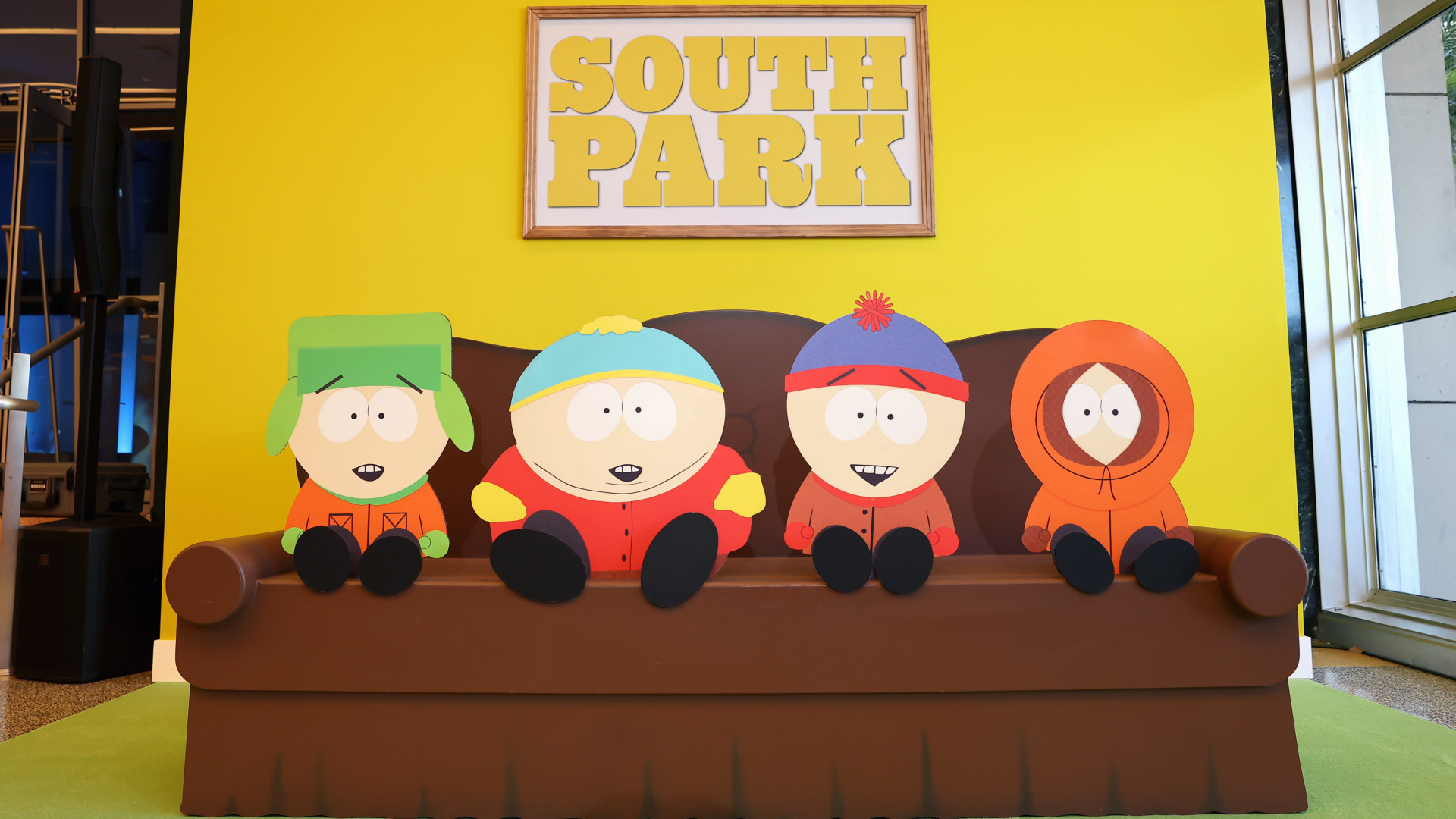 Characters from "South Park" on a couch: Kyle, Cartman, Stan, and Kenny, with a "South Park" sign above them on a yellow wall.