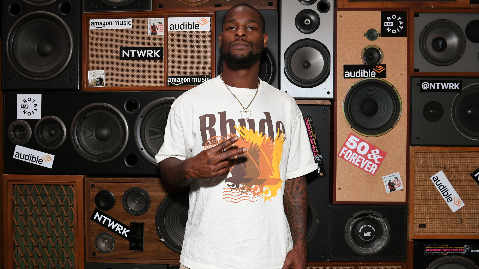 Le'Veon Bell in a white "Rhude" shirt stands in front of a wall of speakers with various brand logos like Amazon Music and Audible.