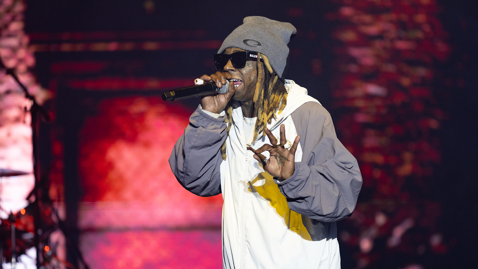 Lil Wayne performing on stage, wearing a gray beanie, sunglasses, and a jacket, holding a microphone.