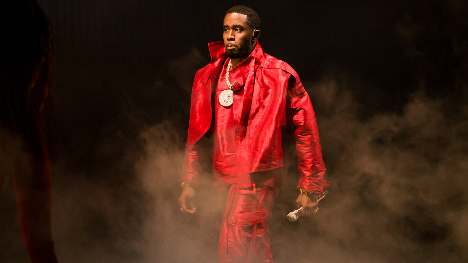 Sean "Diddy" Combs in a red outfit, holding a microphone, with smoke around him on stage.